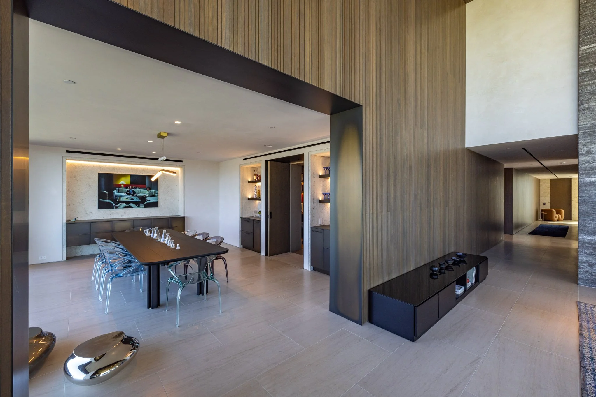 Modern dining room with a long black table, transparent chairs, and a wall-mounted TV. Adjacent to a hallway, featuring built-in shelves with decorative items and a black cabinet.