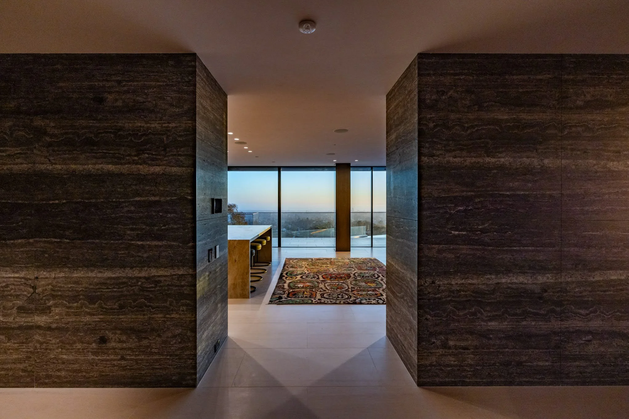 Interior view of a modern room with glass sliding doors revealing a cityscape, a patterned rug on light-colored tile floor, a bar with stools, and dark wood-paneled walls.