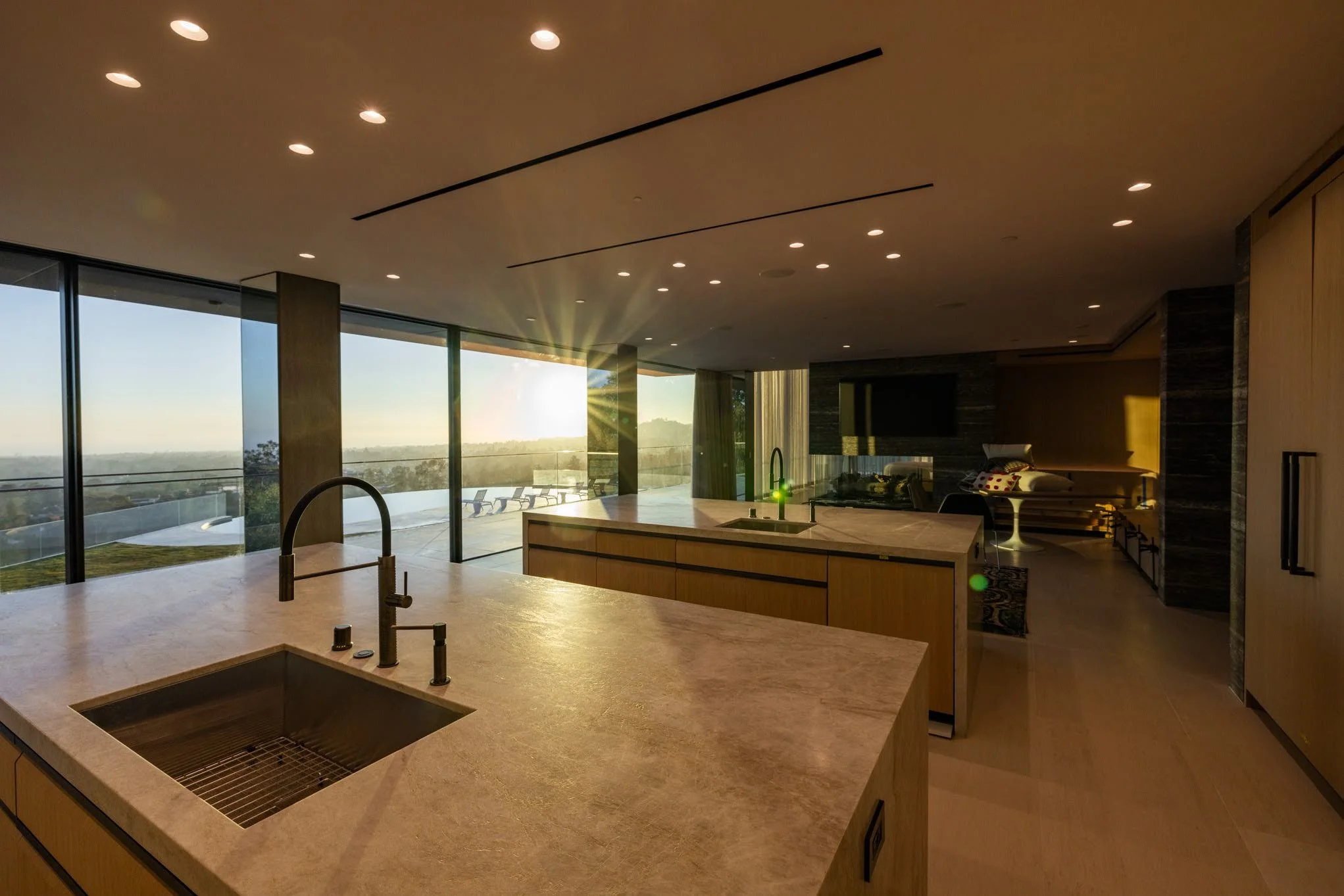 Modern kitchen with a marble countertop island, sink, and a view of a scenic landscape through large glass windows, during sunset.