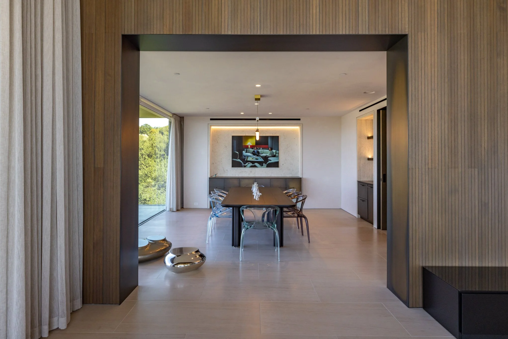 Modern dining room with wooden floors, a rectangular dining table with transparent chairs, a painting on the back wall, and sliding glass doors showing trees outside.