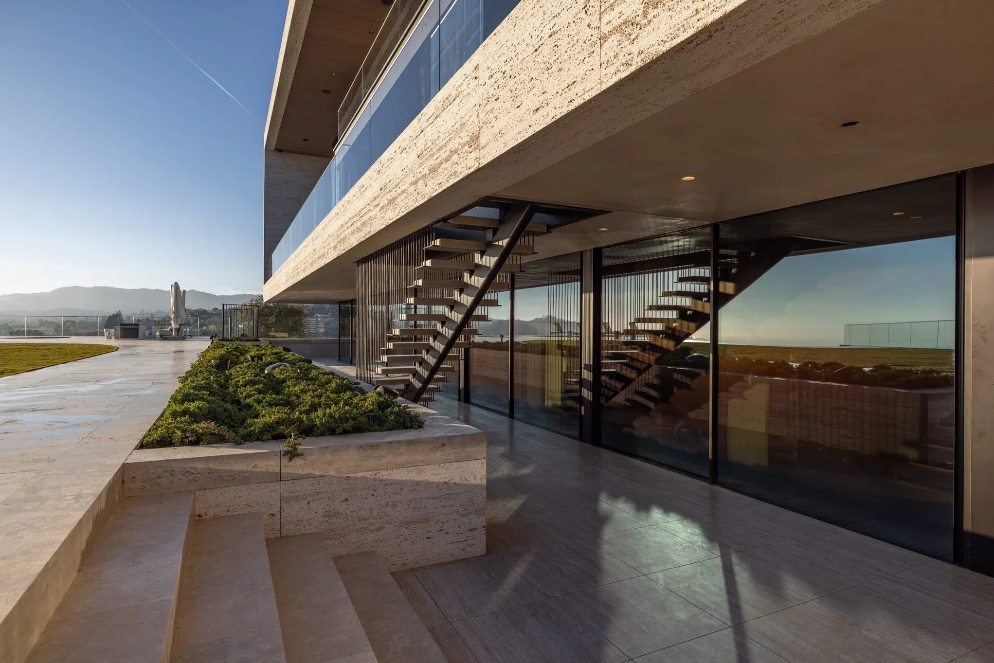 Modern building with glass walls, outdoor stairs, and a landscaped garden with hills and open sky in the background