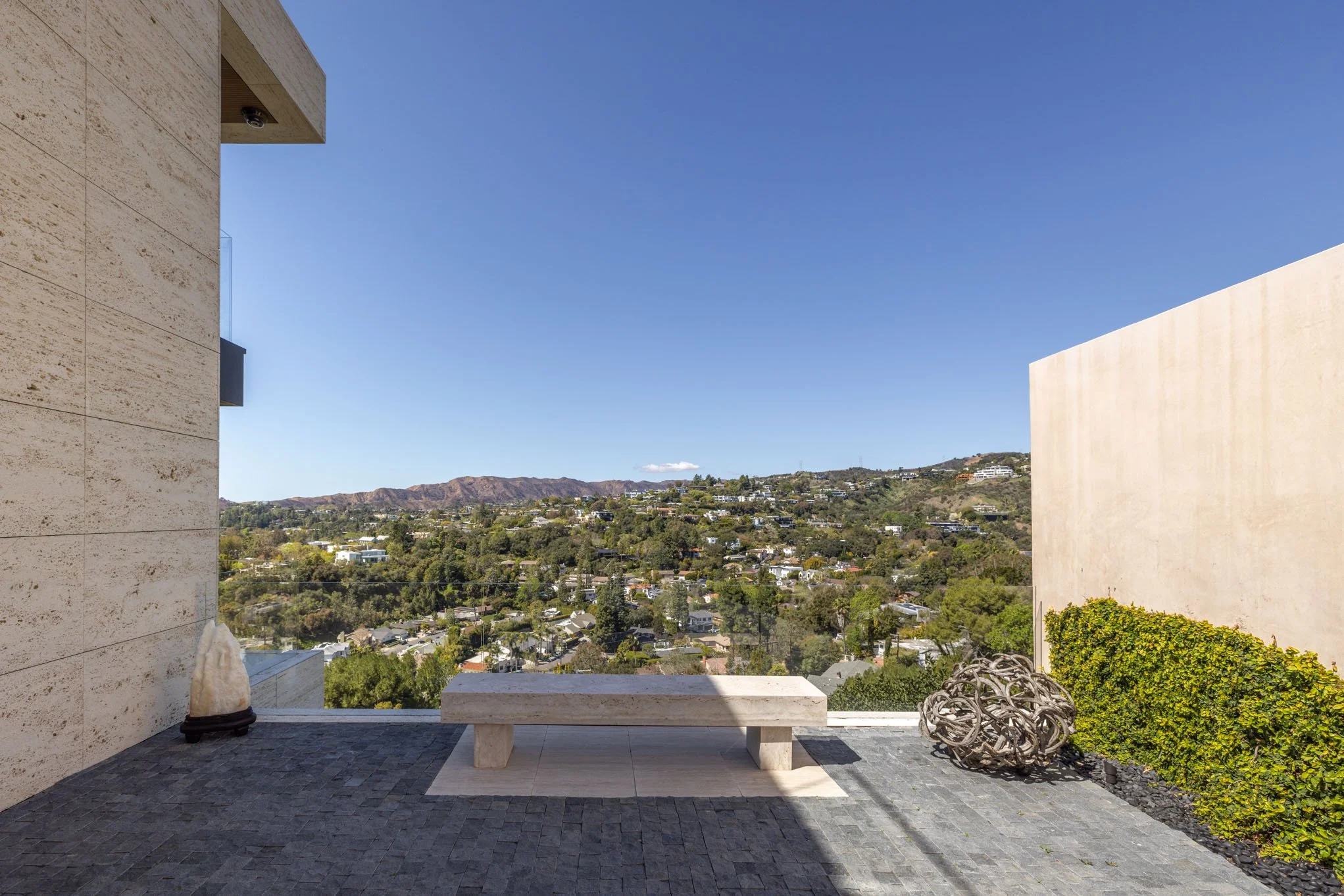 Modern outdoor balcony with bench, decorative sculpture, and a view of a hilly residential neighborhood under a clear blue sky.