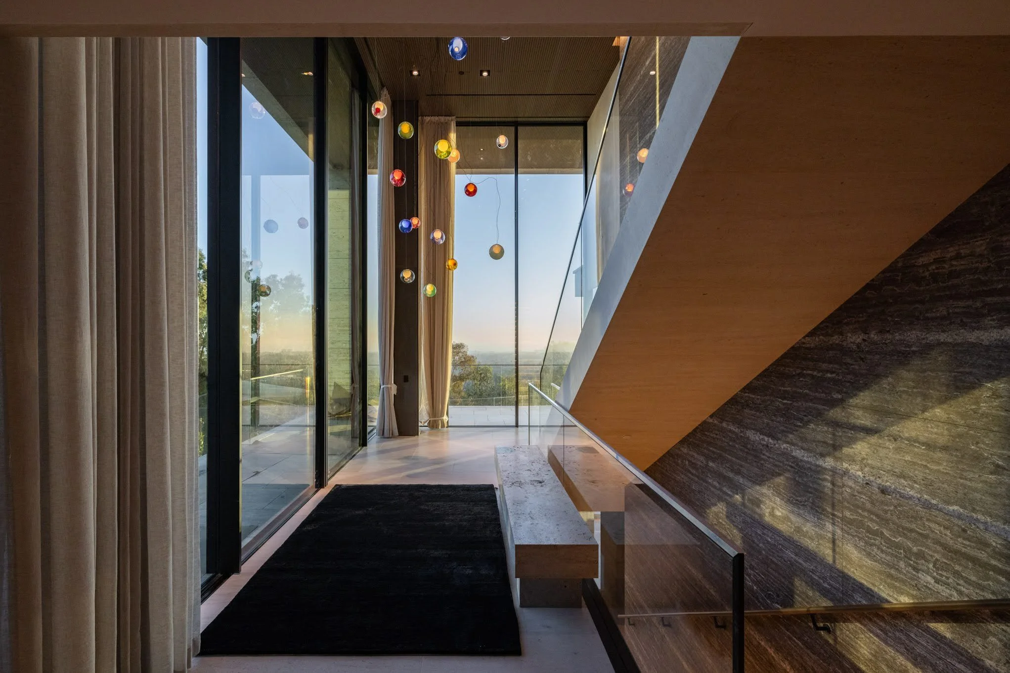 Modern interior with large glass doors, curtains, a black rug, and a colorful chandelier hanging from the ceiling, overlooking a scenic view outside.