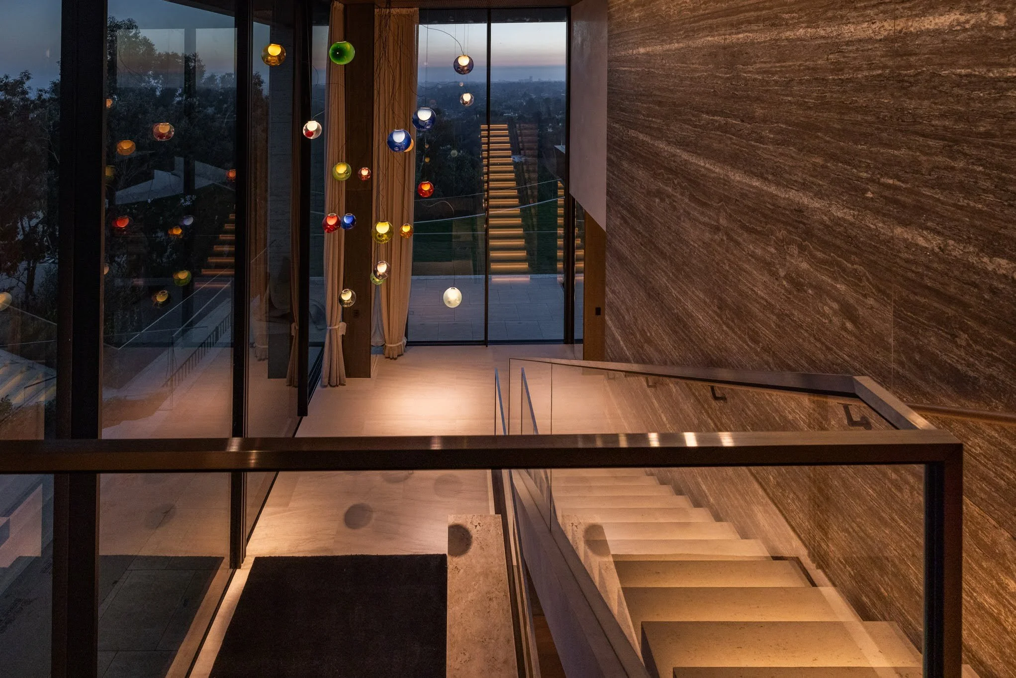 Inside a modern home with a staircase, large glass doors, and hanging colorful glass light fixtures at dusk.