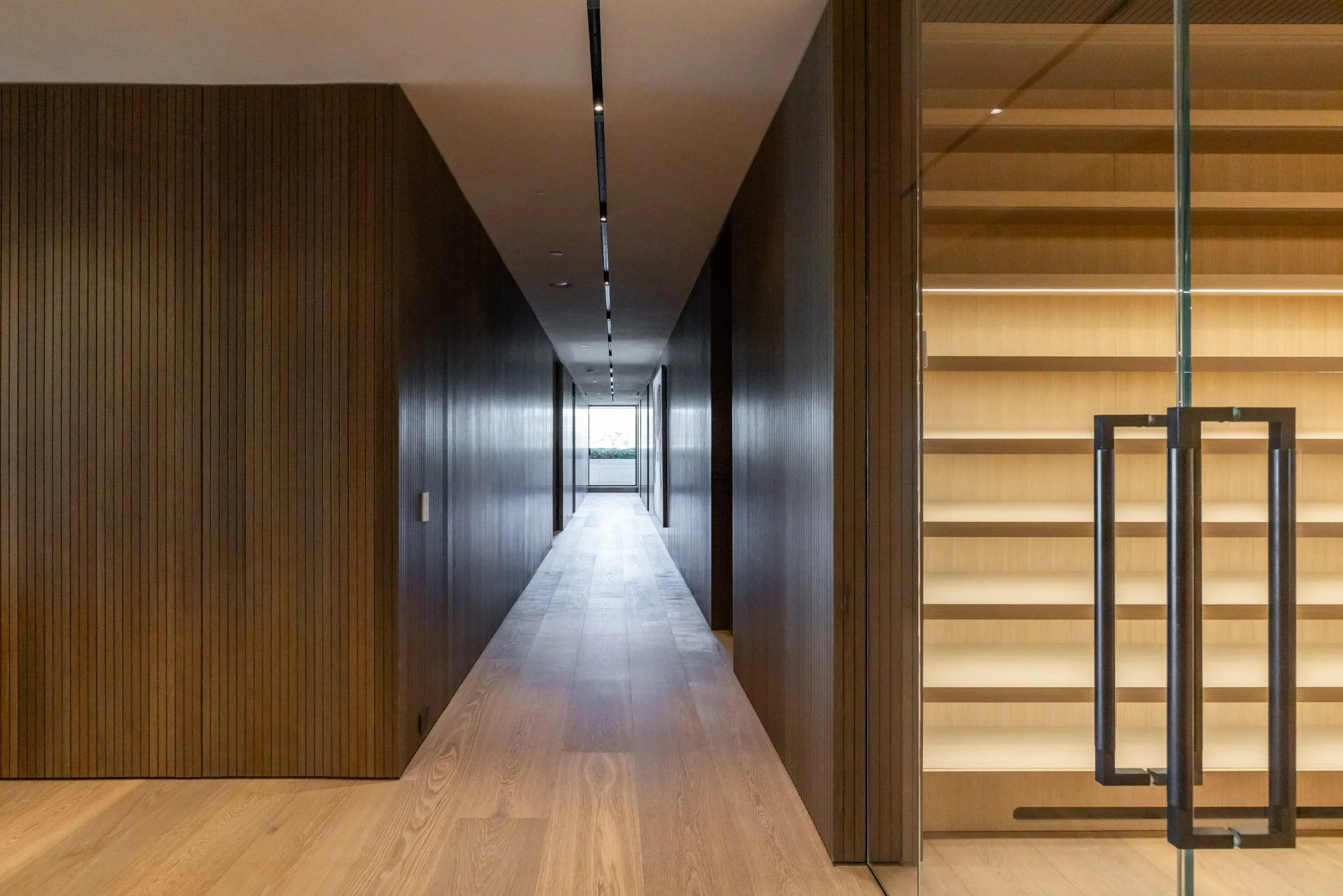 Empty wooden hallway with a door at the end, and a glass door with black handles to the right showing shelves inside.