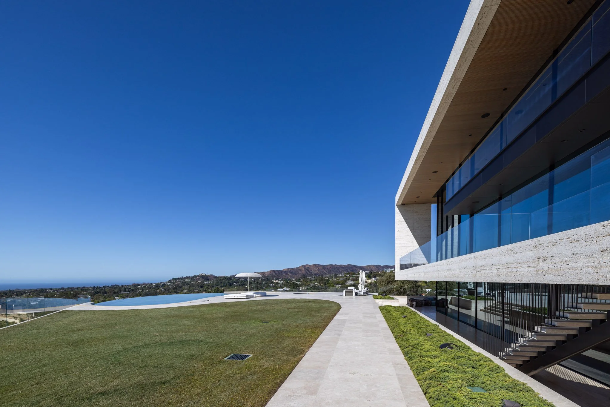 Modern house with large glass windows, a grassy lawn, and an outdoor pool area under a clear blue sky, overlooking distant hills.