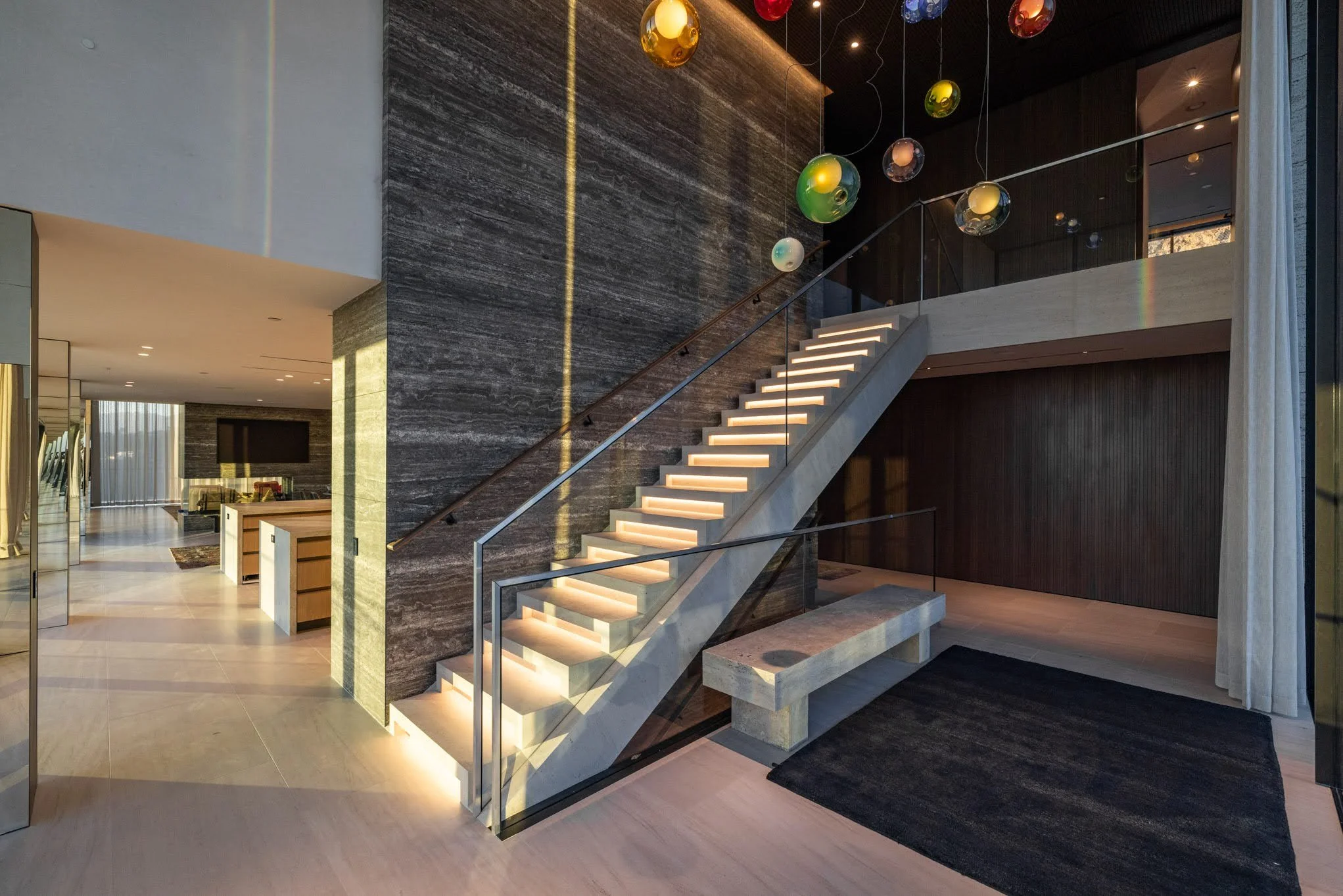 Interior of a modern home with illuminated staircase, dark wood accent wall, colorful hanging spherical pendant lights, and a black rug on a light wooden floor.
