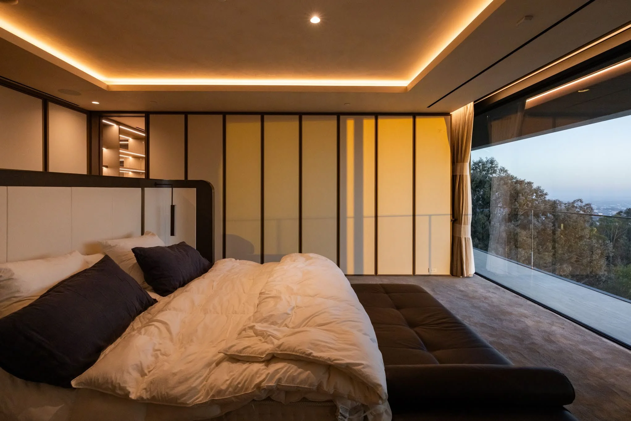 Modern bedroom with a large bed, dark pillows, a beige headboard, wood-paneled walls, and a floor-to-ceiling window showing trees and an outdoor view during dusk.