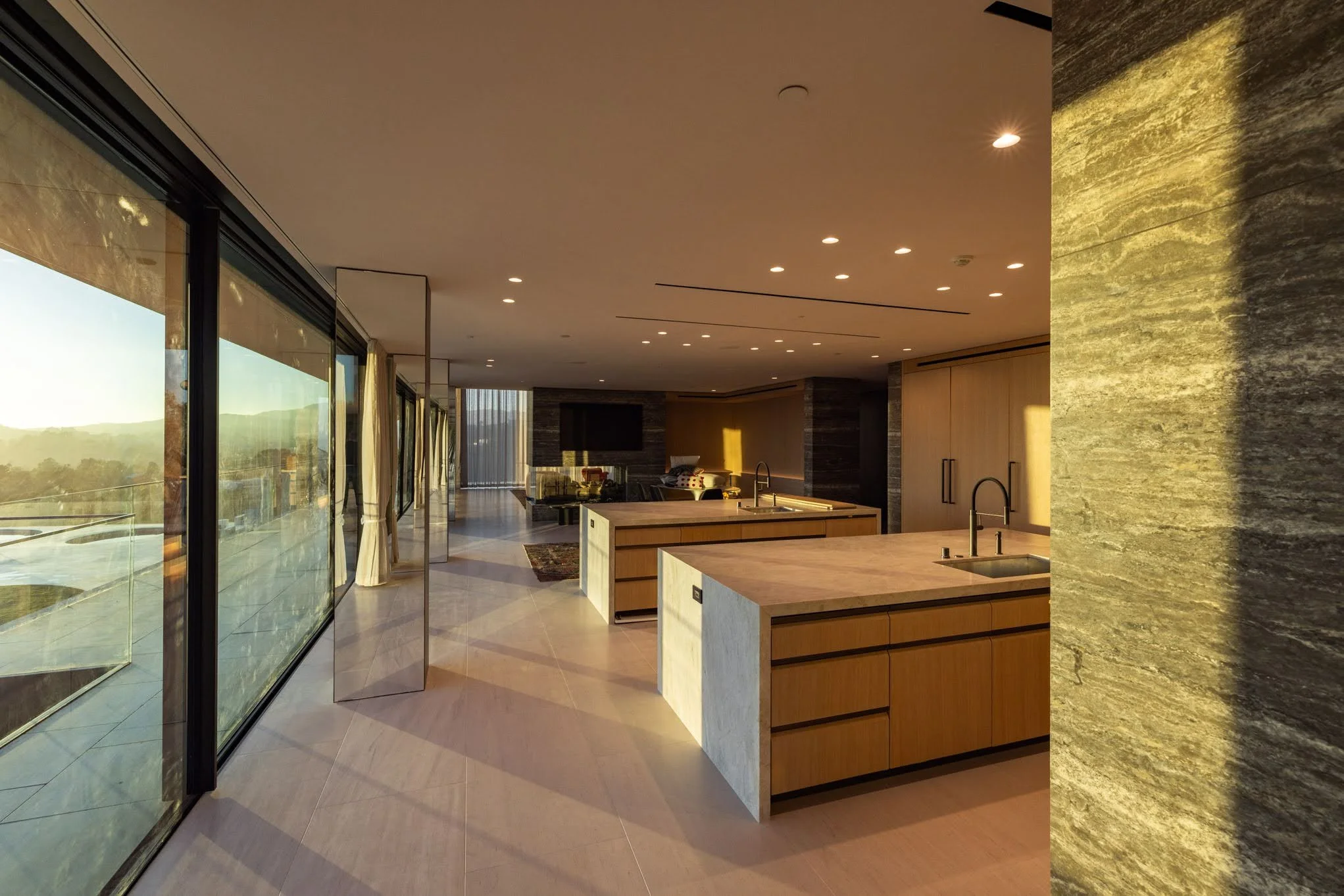 Modern open-concept kitchen with wooden cabinetry, central island with double sinks, large glass windows with curtains, and a view of a living area with couches and a wall-mounted TV, illuminated by recessed ceiling lights, during sunset.