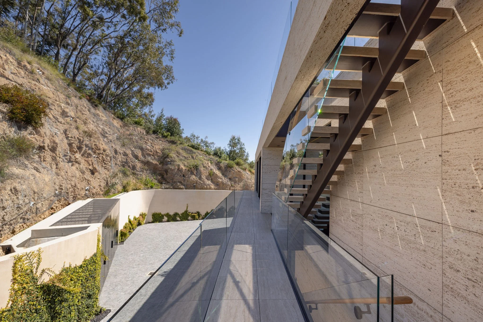 Modern building exterior with glass balcony, outdoor staircase, beige stone walls, and a hillside with trees in the background under a clear blue sky.