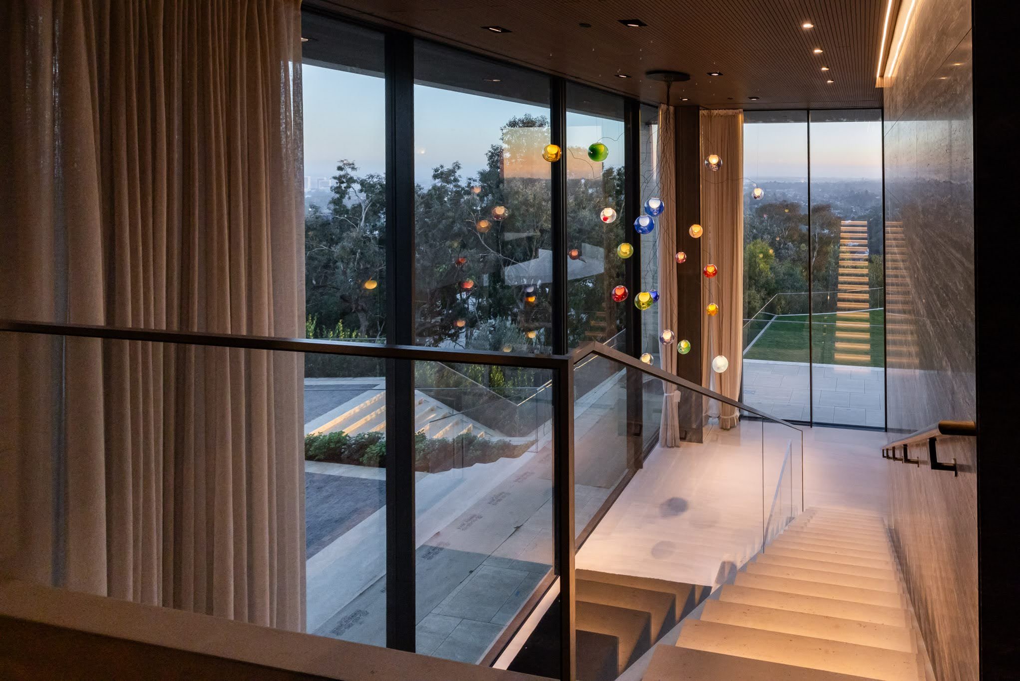 Modern staircase with large glass windows and a colorful hanging light fixture in a contemporary building with a view of trees and a distant cityscape.
