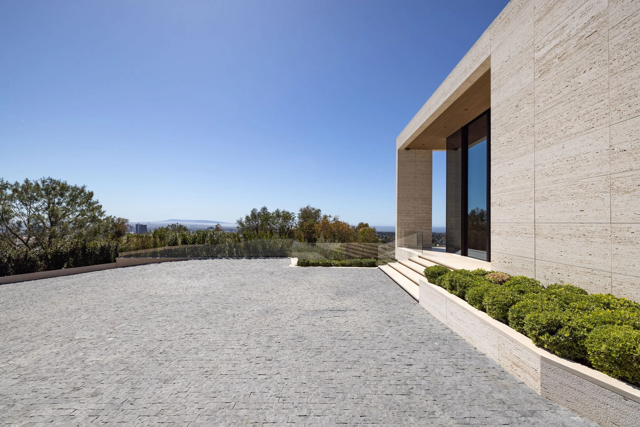 Modern beige stone house with glass door and steps, landscaped with green bushes, overlooking a clear blue sky and distant trees.