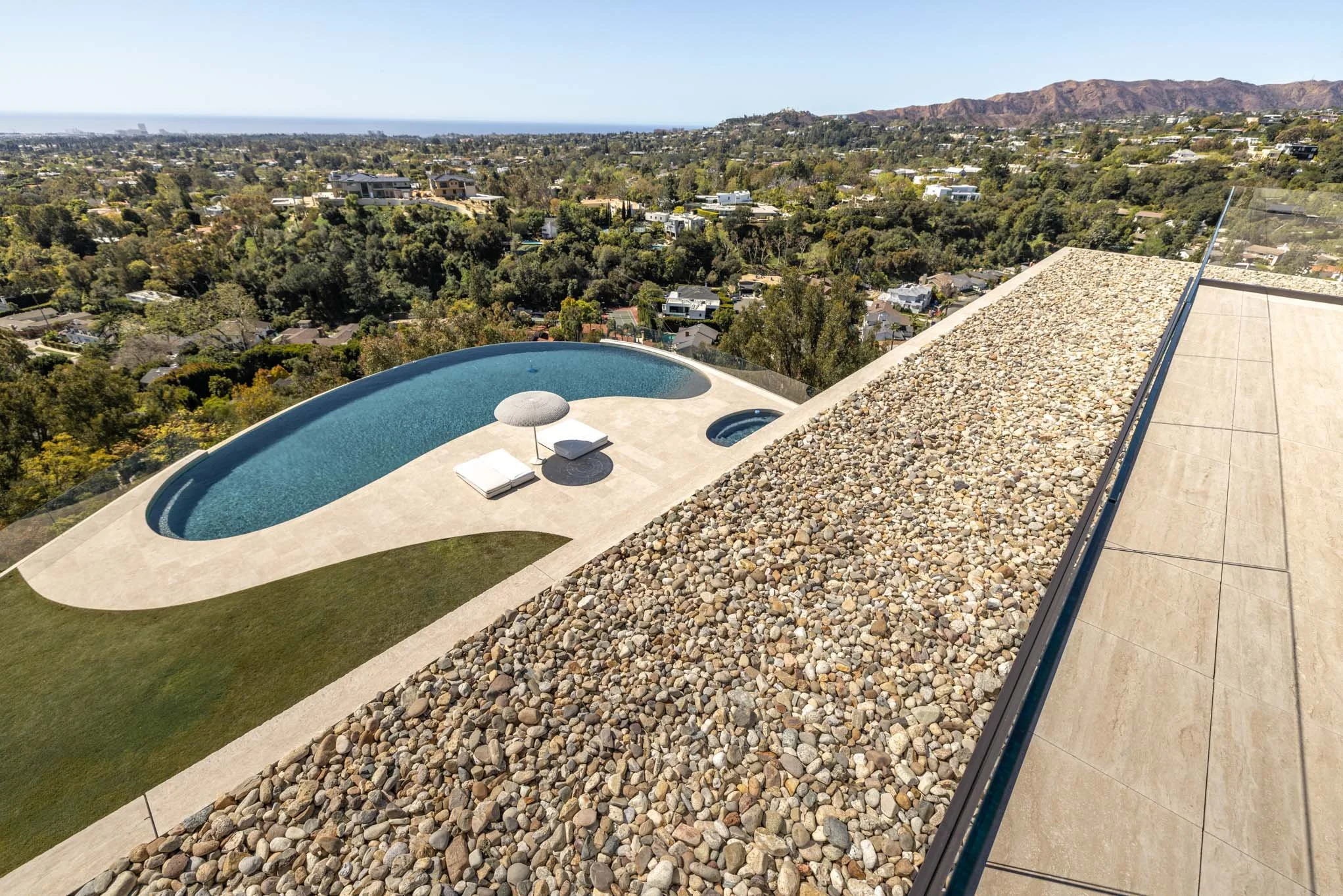 A rooftop with a swimming pool, a poolside umbrella, and lounge chairs under a sunny sky, overlooking a cityscape with hills and mountains in the distance.
