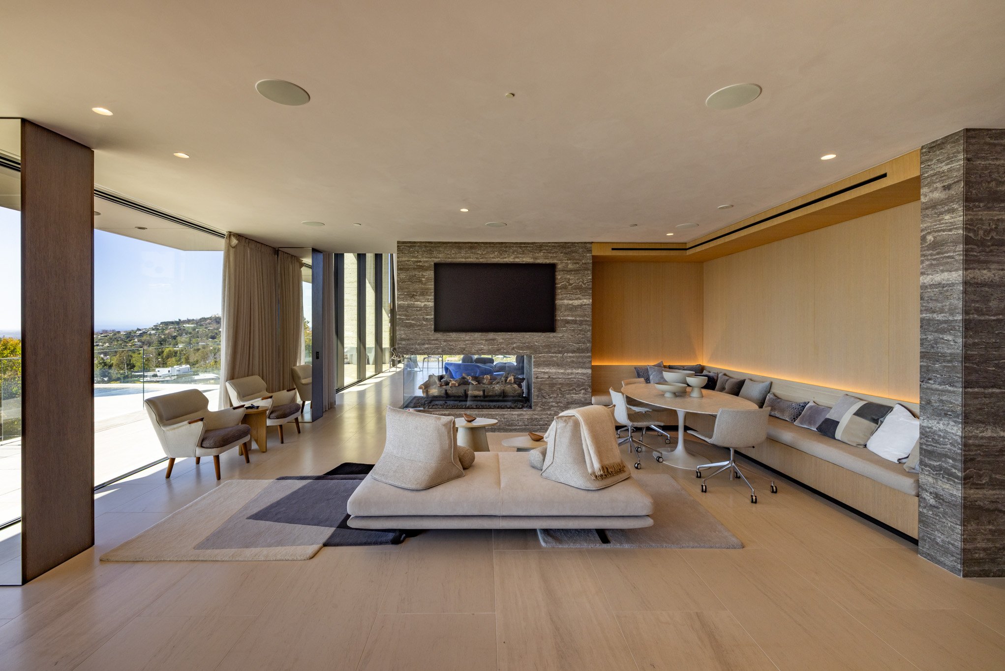 Modern living room with beige sofas, a fireplace, a large flat-screen TV, and a dining area with a long curved table and chairs, illuminated by recessed lighting and large windows showing an outdoor view.