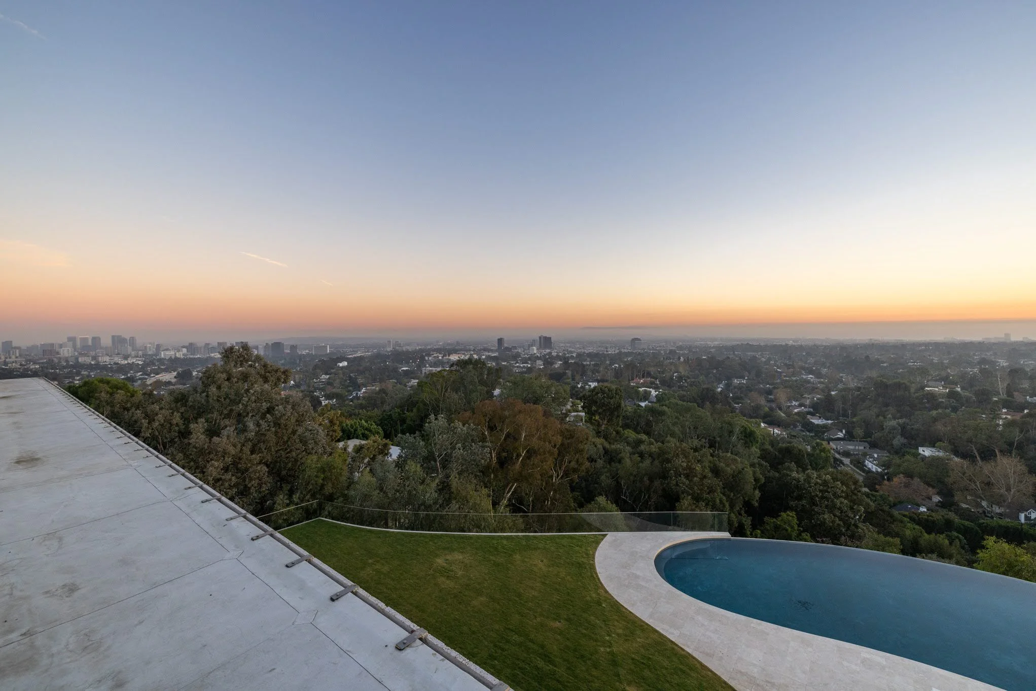 Cityscape view at sunset from a rooftop with a swimming pool and greenery in the foreground.