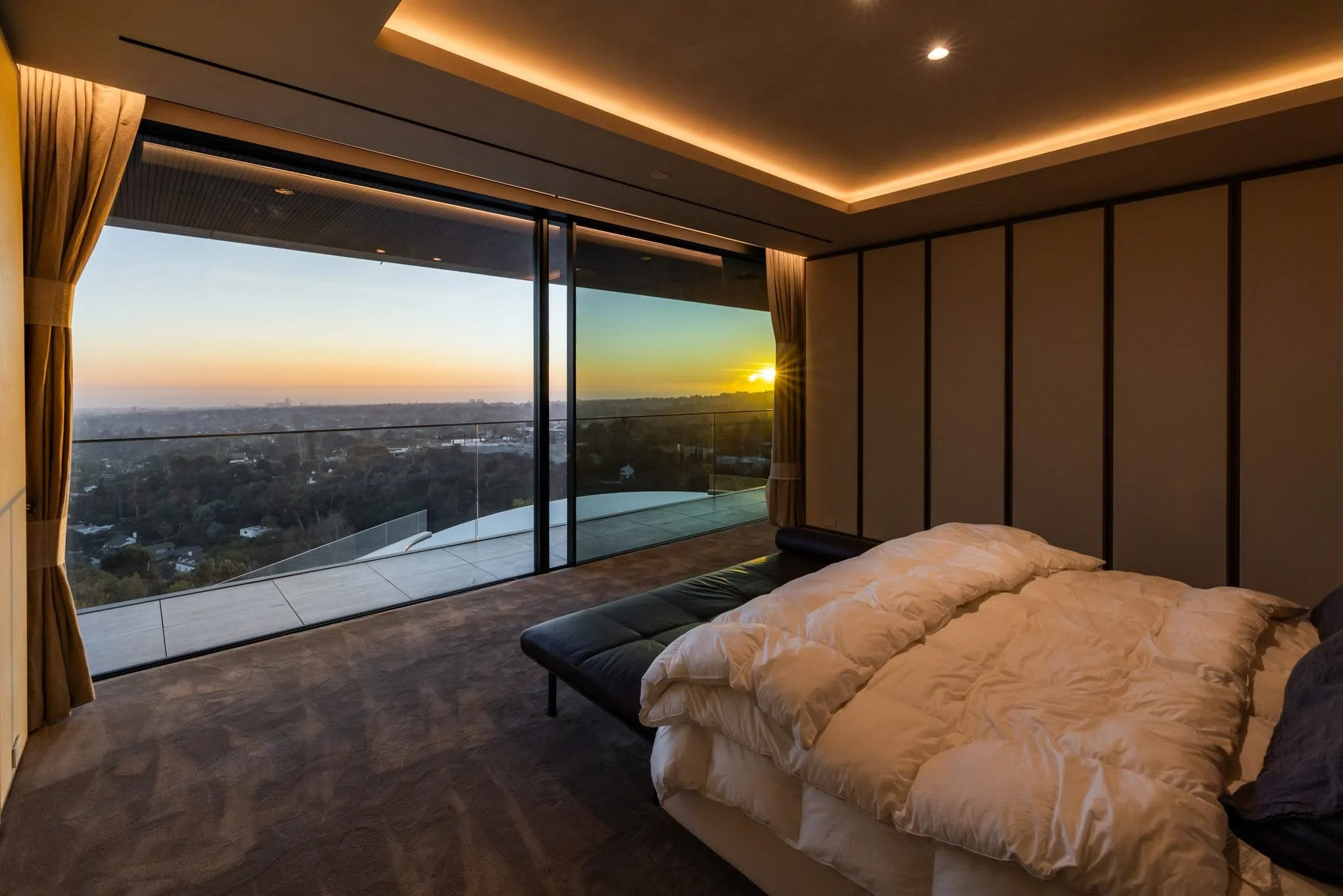 A modern bedroom with a large bed and white bedding, next to a glass wall with a view of a cityscape and sunset, with beige curtains and soft ceiling lighting.