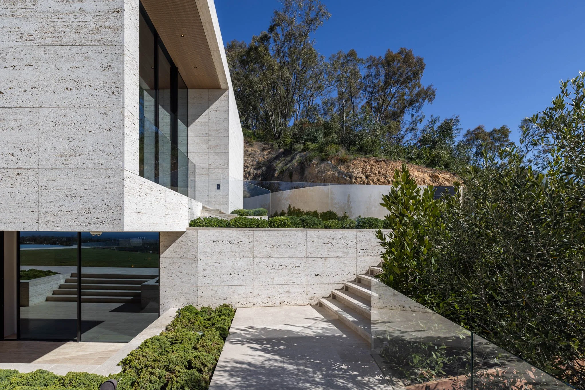Modern house with large glass windows, concrete walls, stairs, green shrubs, and trees under a blue sky.