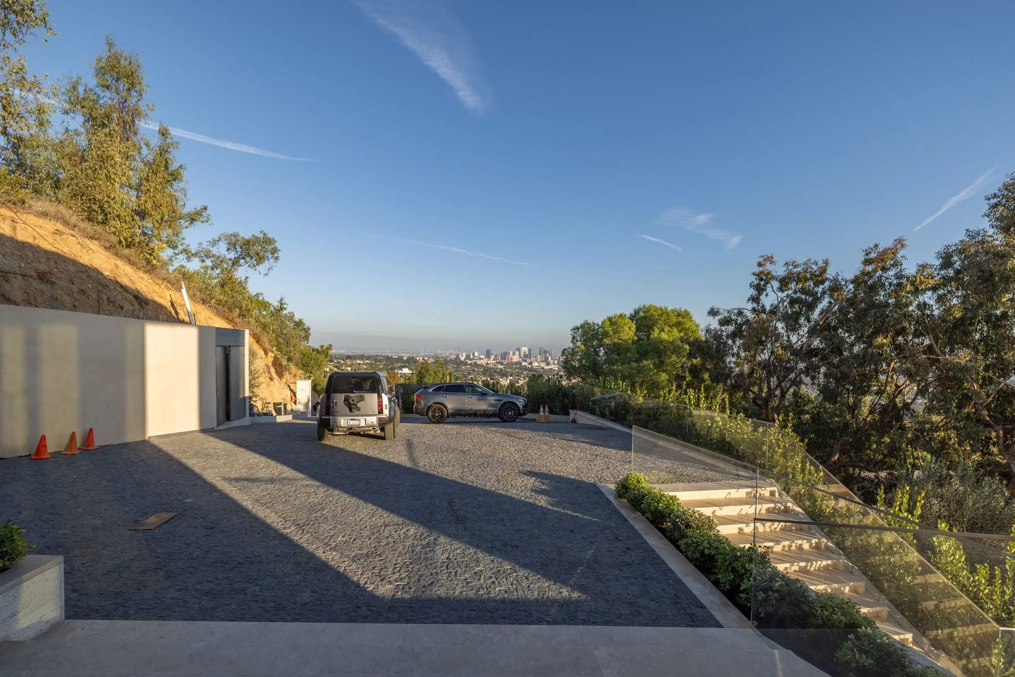 Parking lot with three cars, granite pavement, overlooking a city skyline in the distance, surrounded by trees and hillside, during late afternoon sunlight.