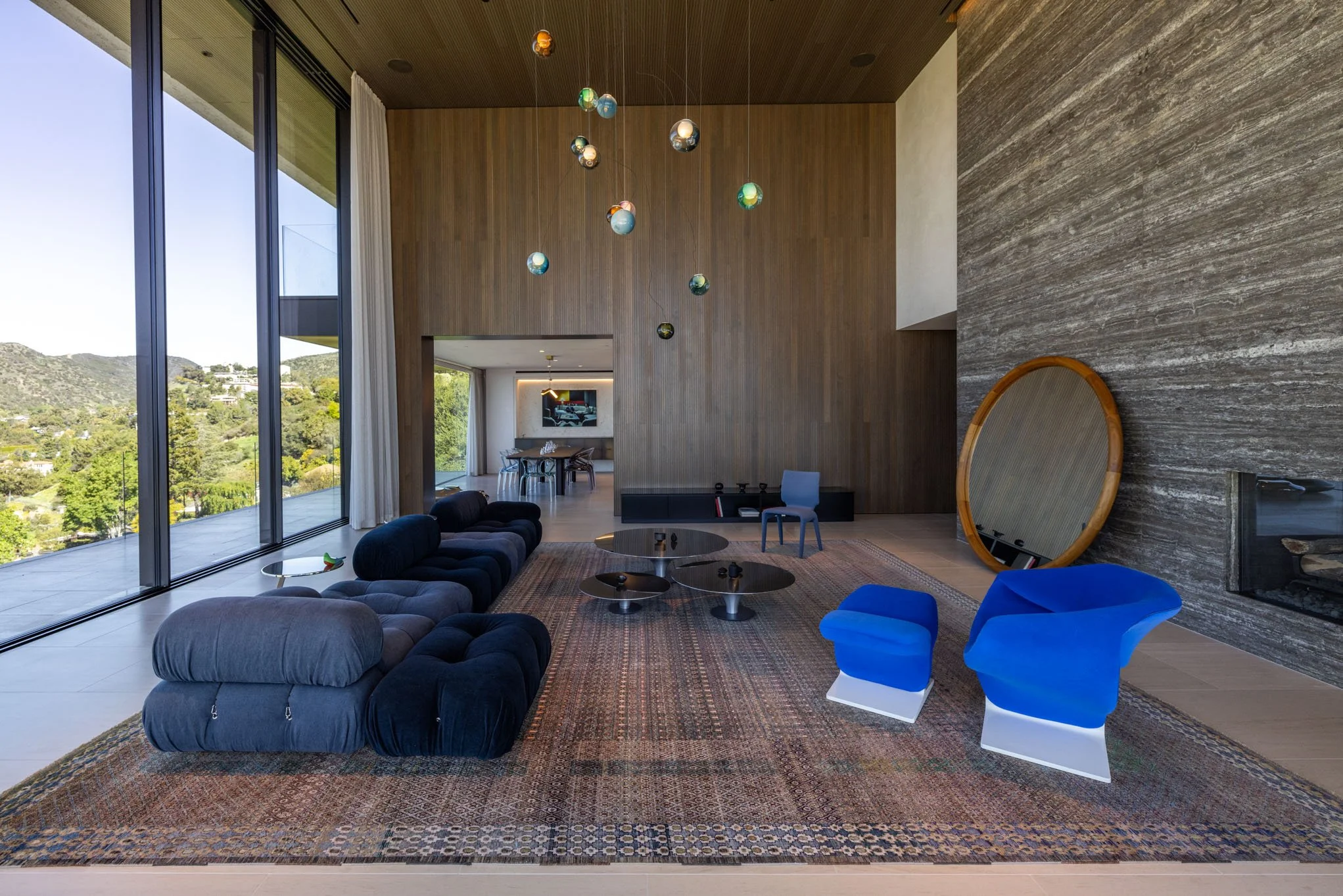 Modern living room with large floor-to-ceiling windows, featuring a black sofa, blue armchairs, a large round mirror, a colorful patterned rug, and a dining area in the background.