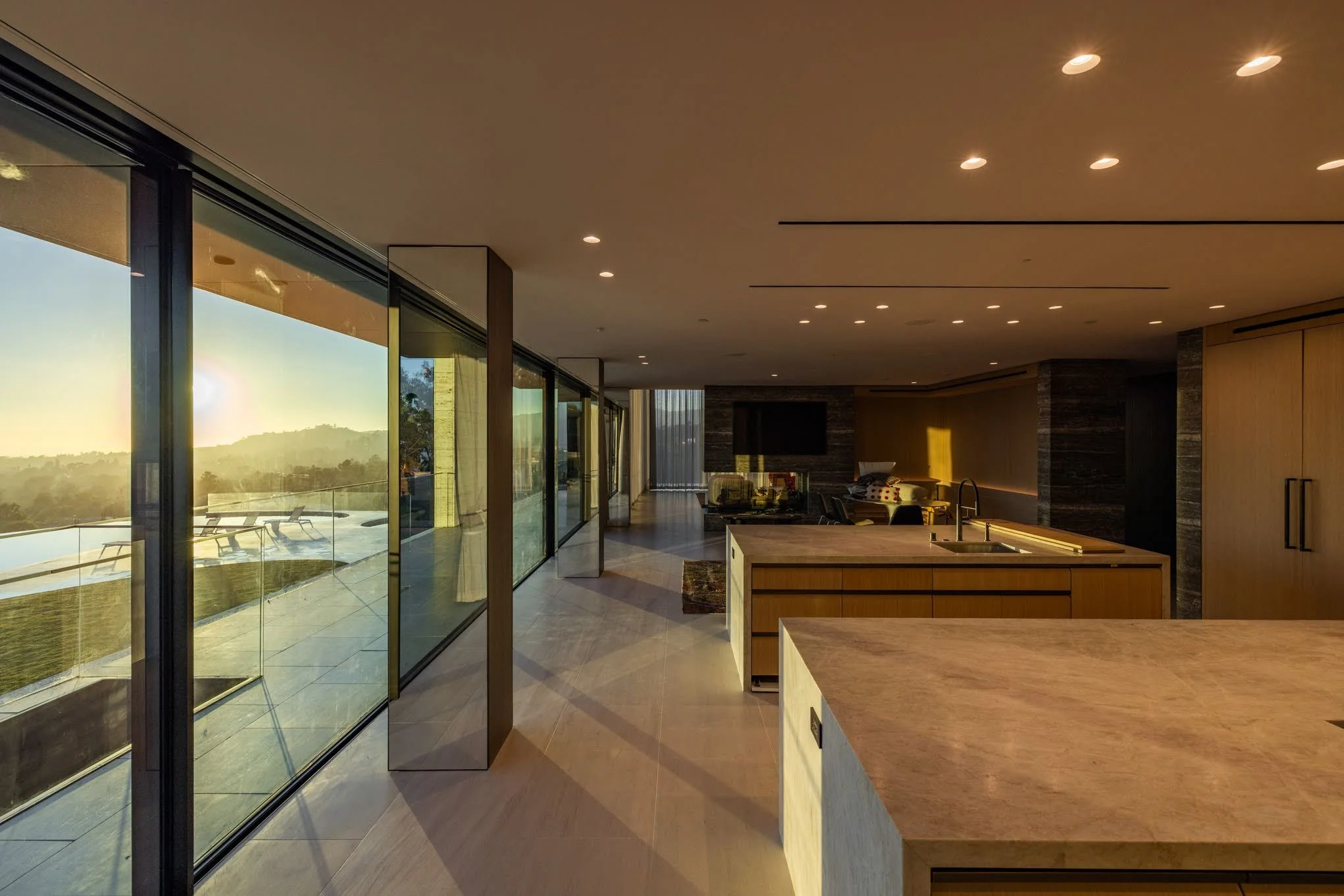 Modern kitchen and living area with large glass windows showing an outdoor deck and a sunset view of hills.