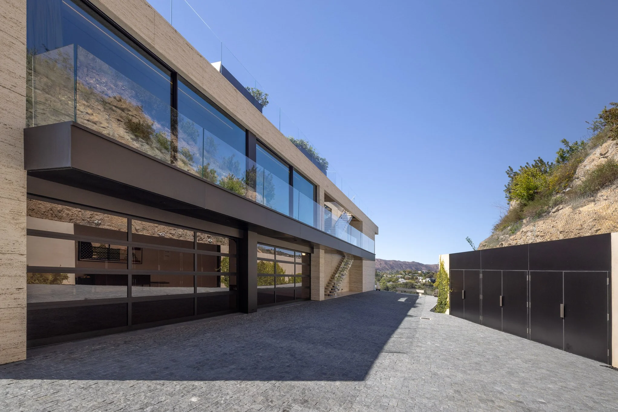 Modern multi-story building with glass balconies and a garage door, located next to a hillside under a clear blue sky.