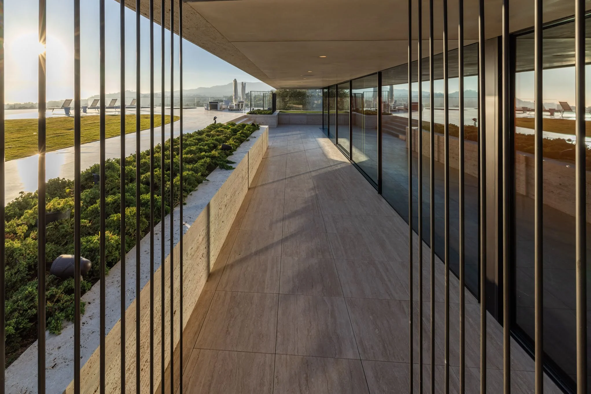Modern balcony with glass sliding doors, metal bars, potted plants, and a view of a pool area, lounge chairs, water, and mountains in the distance.