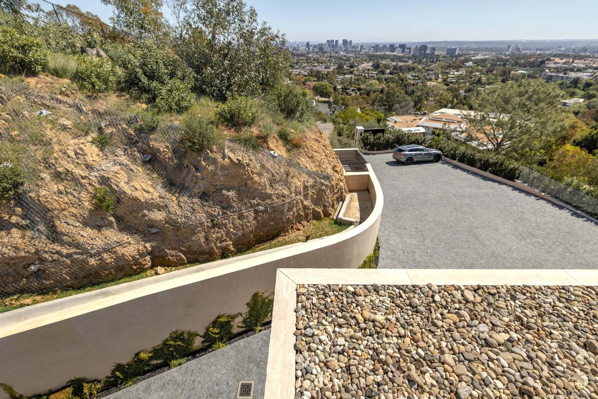 Top view of a parking area on a hillside with a black car, stone and gravel surfaces, a curved white wall, and a city skyline in the distance.