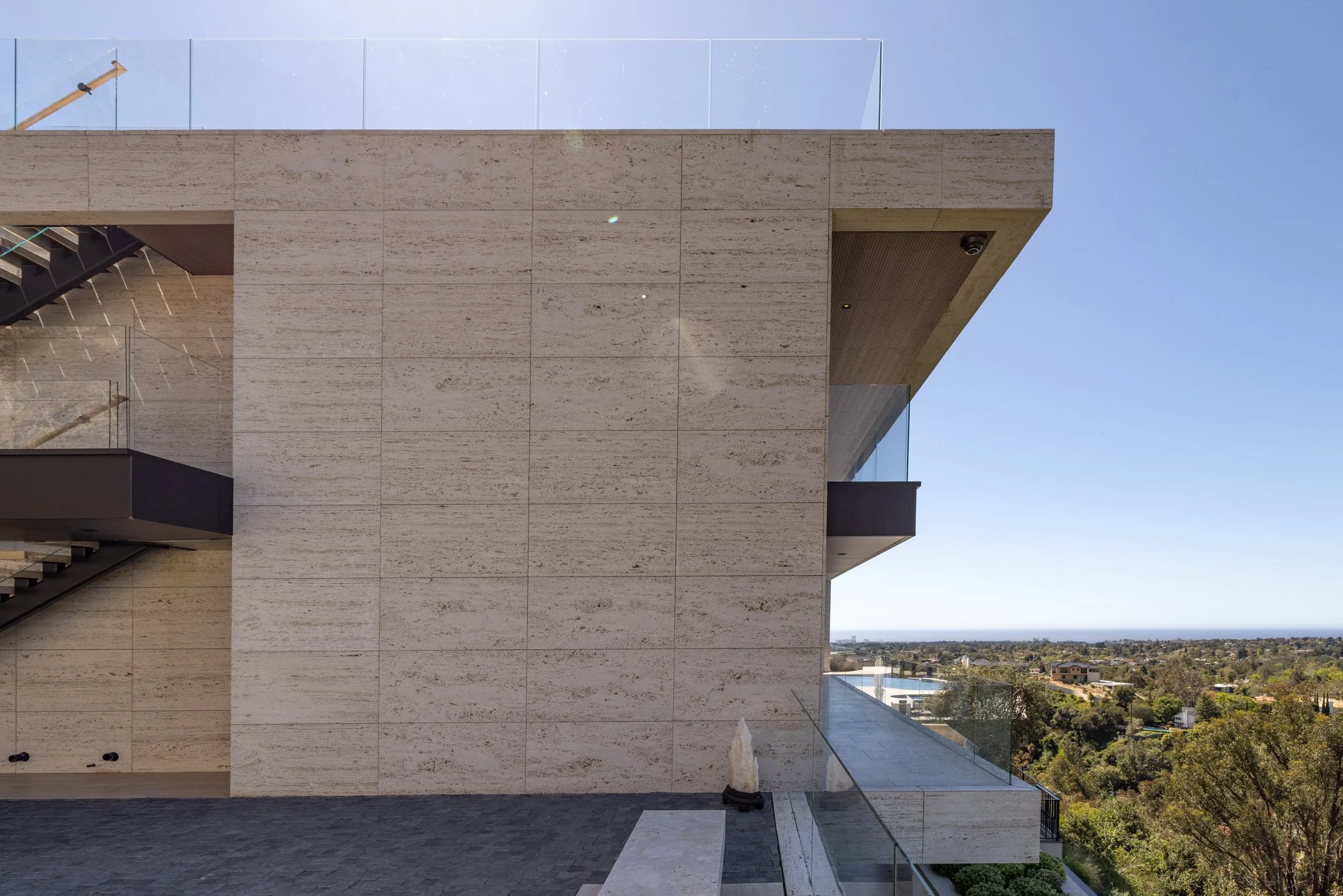 Modern building with beige stone exterior, glass railing, and outdoor staircase, overlooking a scenic landscape with trees and distant horizon.