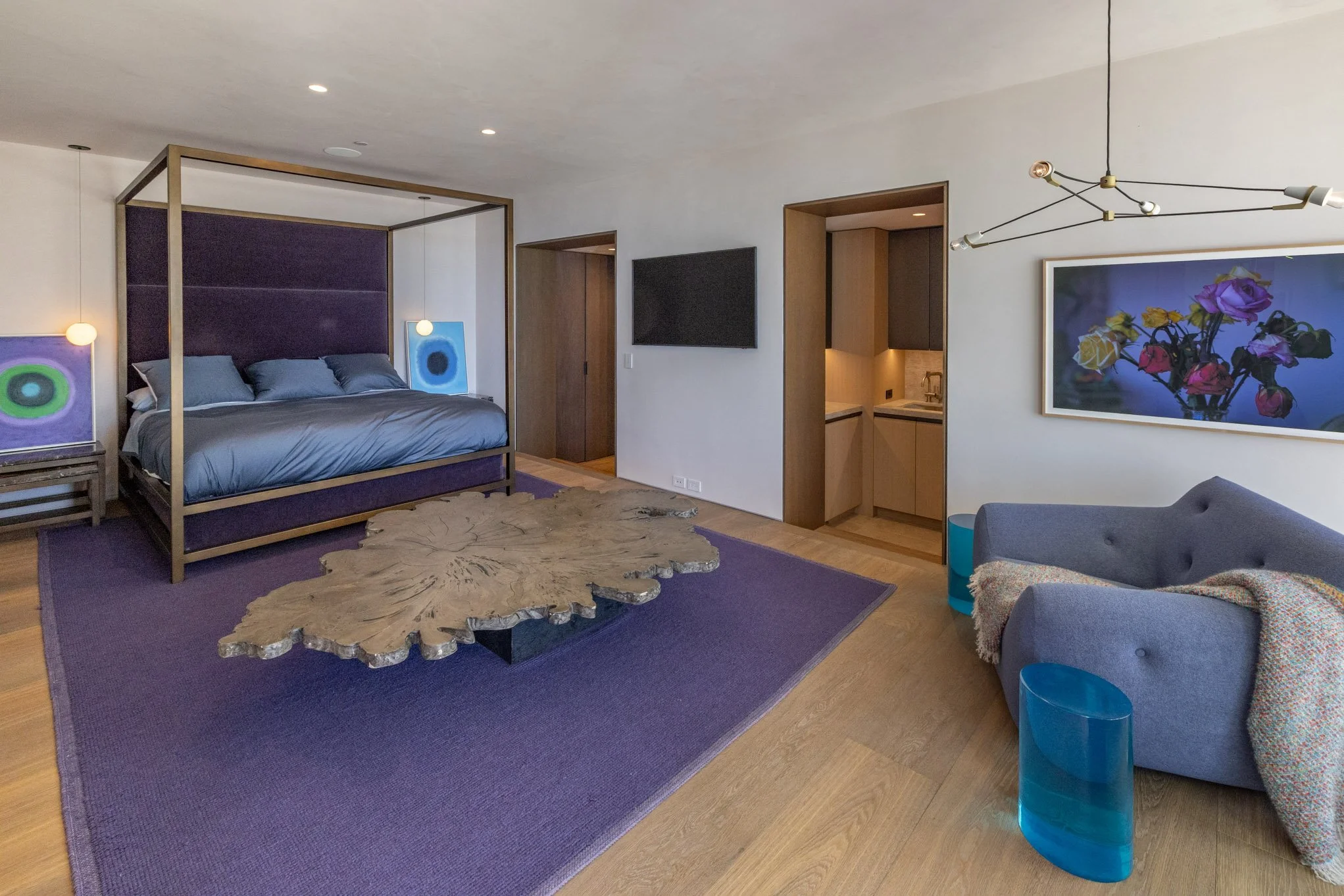 Modern bedroom with four-poster bed, purple rug, wooden coffee table, gray armchair with colorful blanket, artwork, and a small kitchenette with a wall-mounted TV.