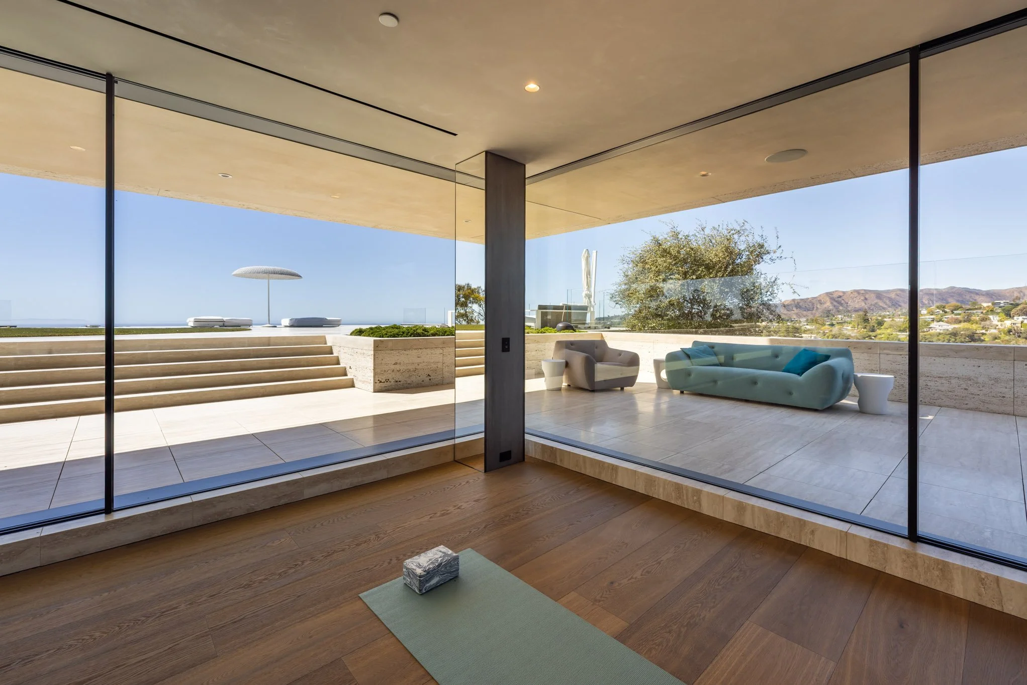 Modern living space with large glass sliding doors overlooking outdoor patio with steps, lounge chairs, and a beach umbrella, with scenic hills and trees in the background.