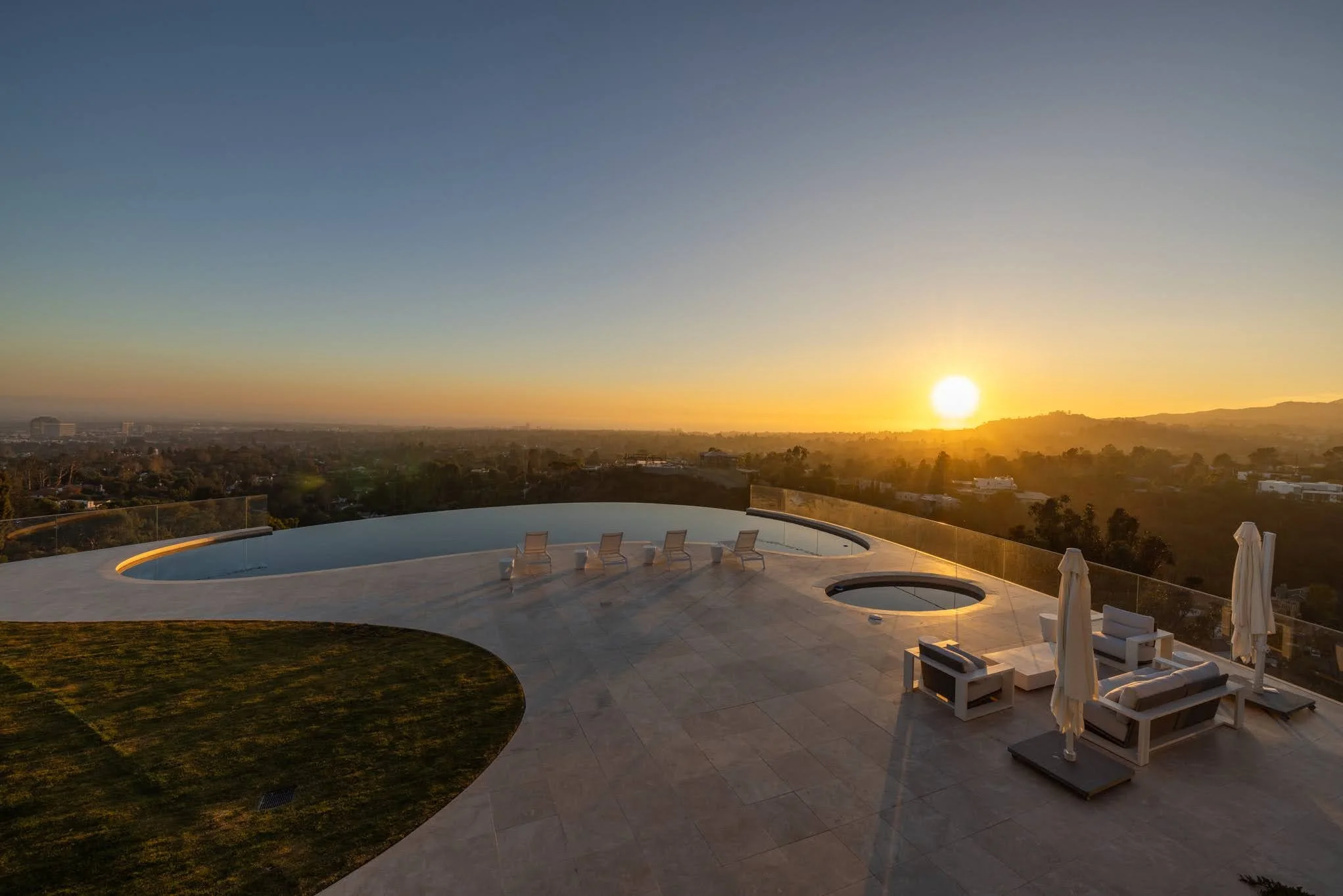 Sunset view from an infinity pool area with lounge chairs, umbrellas, and a hot tub, overlooking a distant landscape.