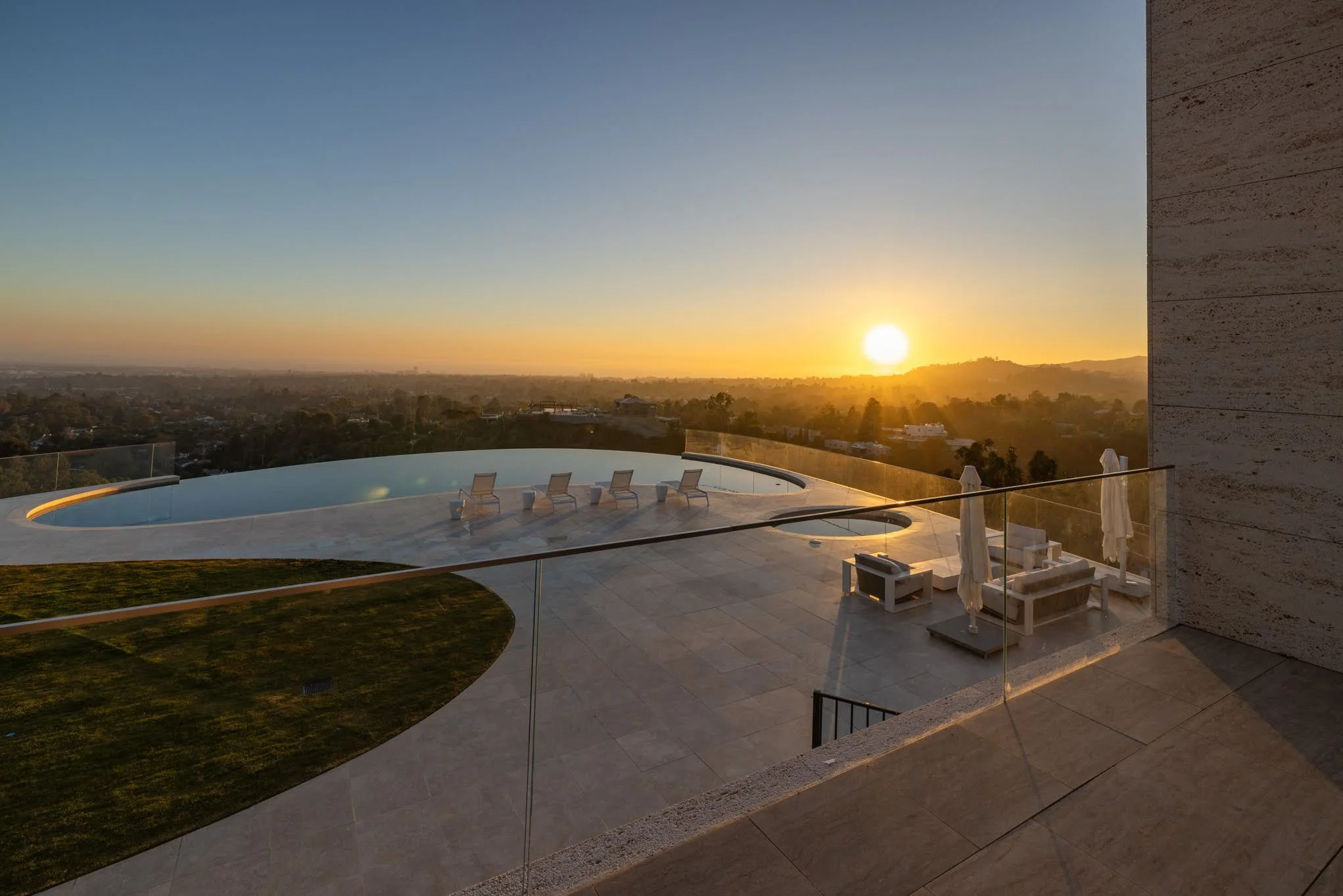 Sunset view from a rooftop balcony with lounge chairs, outdoor seating area with umbrellas, and an infinity pool overlooking a cityscape.