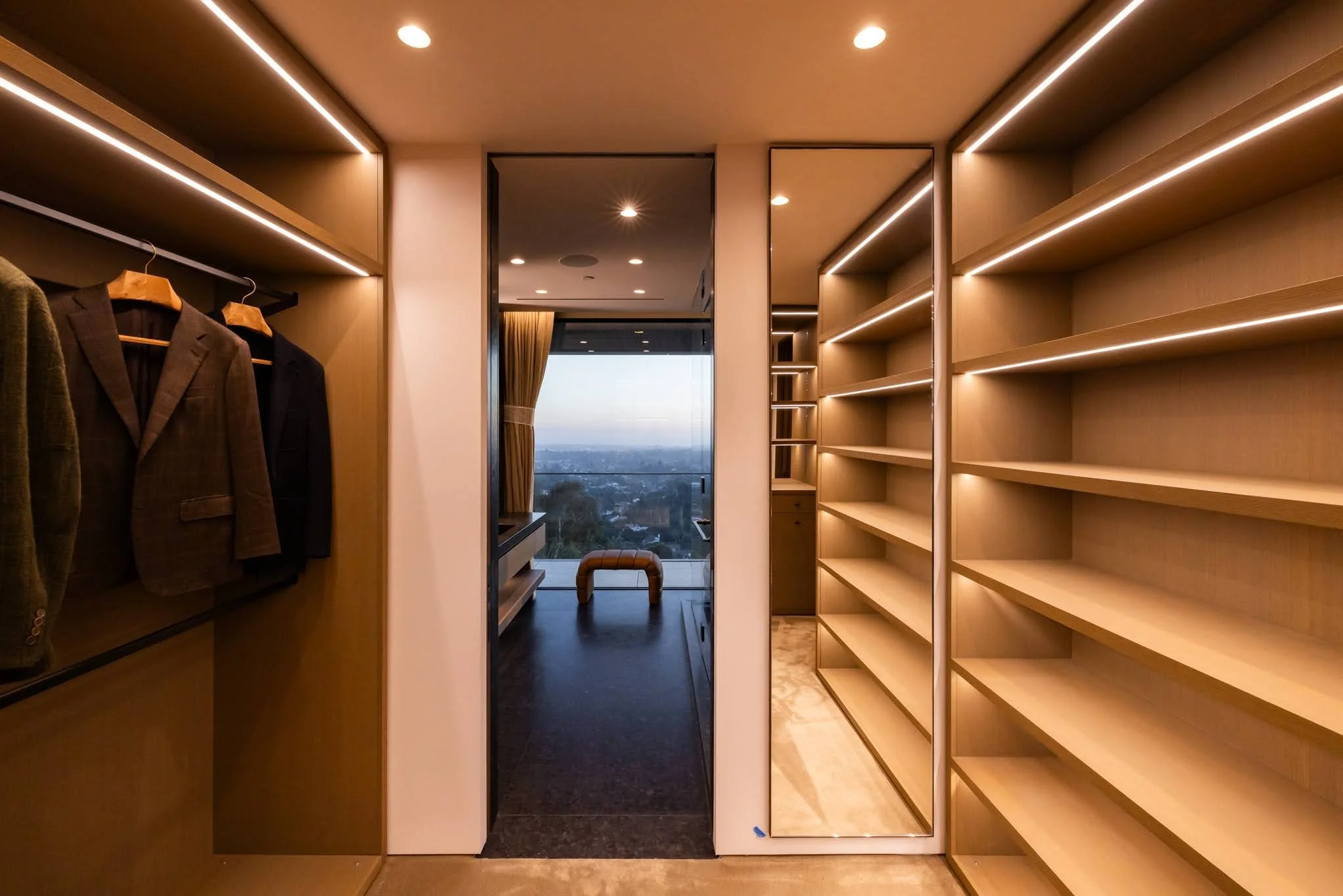 Modern walk-in closet with built-in wooden shelves and hanging space, a large mirror, and a view of a cityscape through a window in the background.