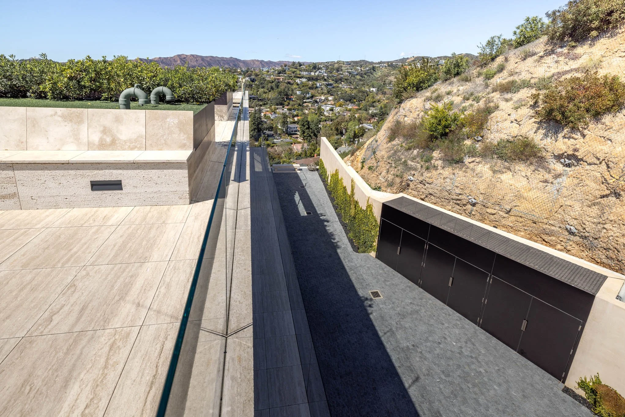 Modern rooftop terrace with beige tiled flooring, glass railing, and a green garden area, overlooking a hillside and cityscape in the distance.