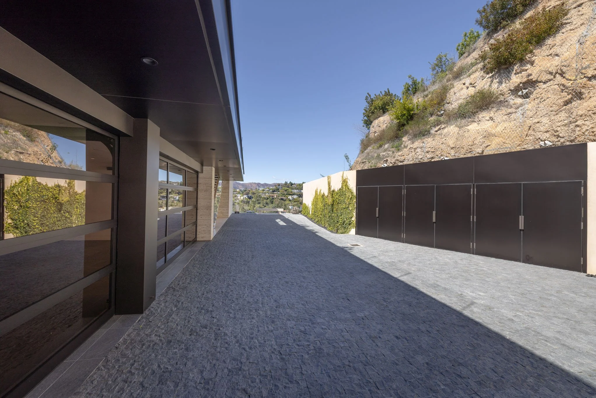 Modern outdoor patio area with dark tiled flooring, large glass windows on the left, and a tall beige wall with black gates on the right, overlooking a hillside with sparse vegetation under a clear blue sky.