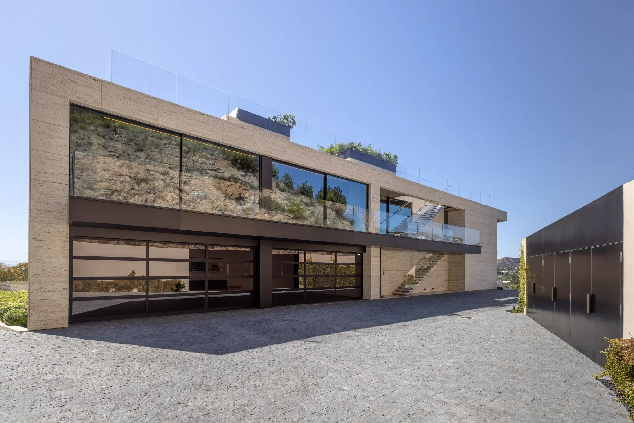 Modern multi-story house with large glass windows, stone exterior, outdoor staircase, rooftop garden with plants, paved driveway, and black gate.
