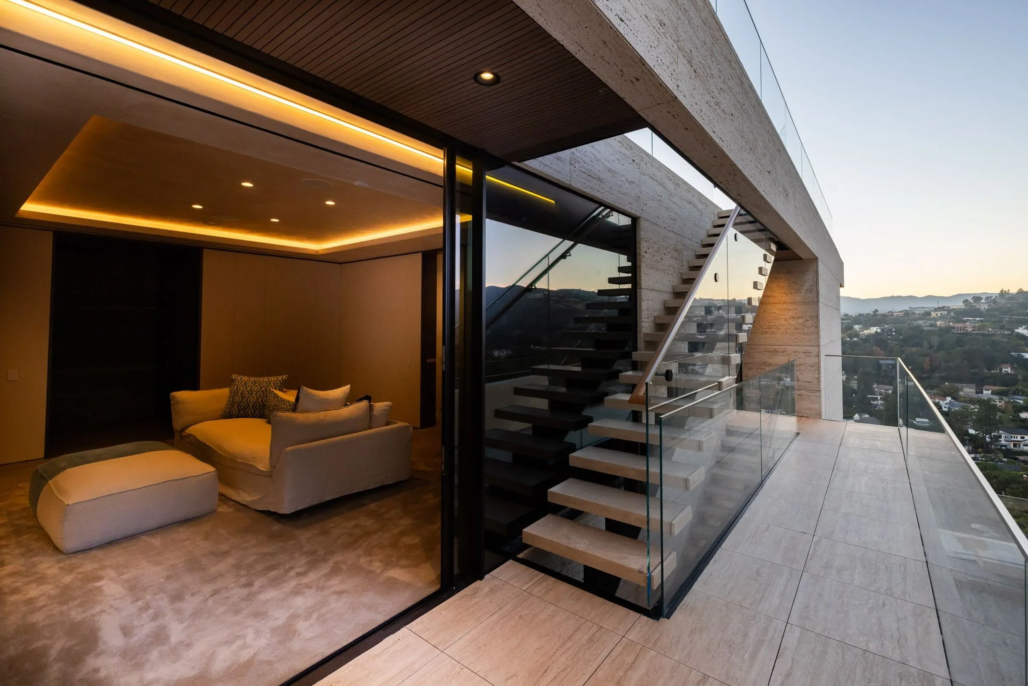 Modern living room with beige sofa, ottoman, and decorative pillows, large glass sliding door leading to an outdoor terrace with a staircase, overlooking a cityscape and hills at sunset.