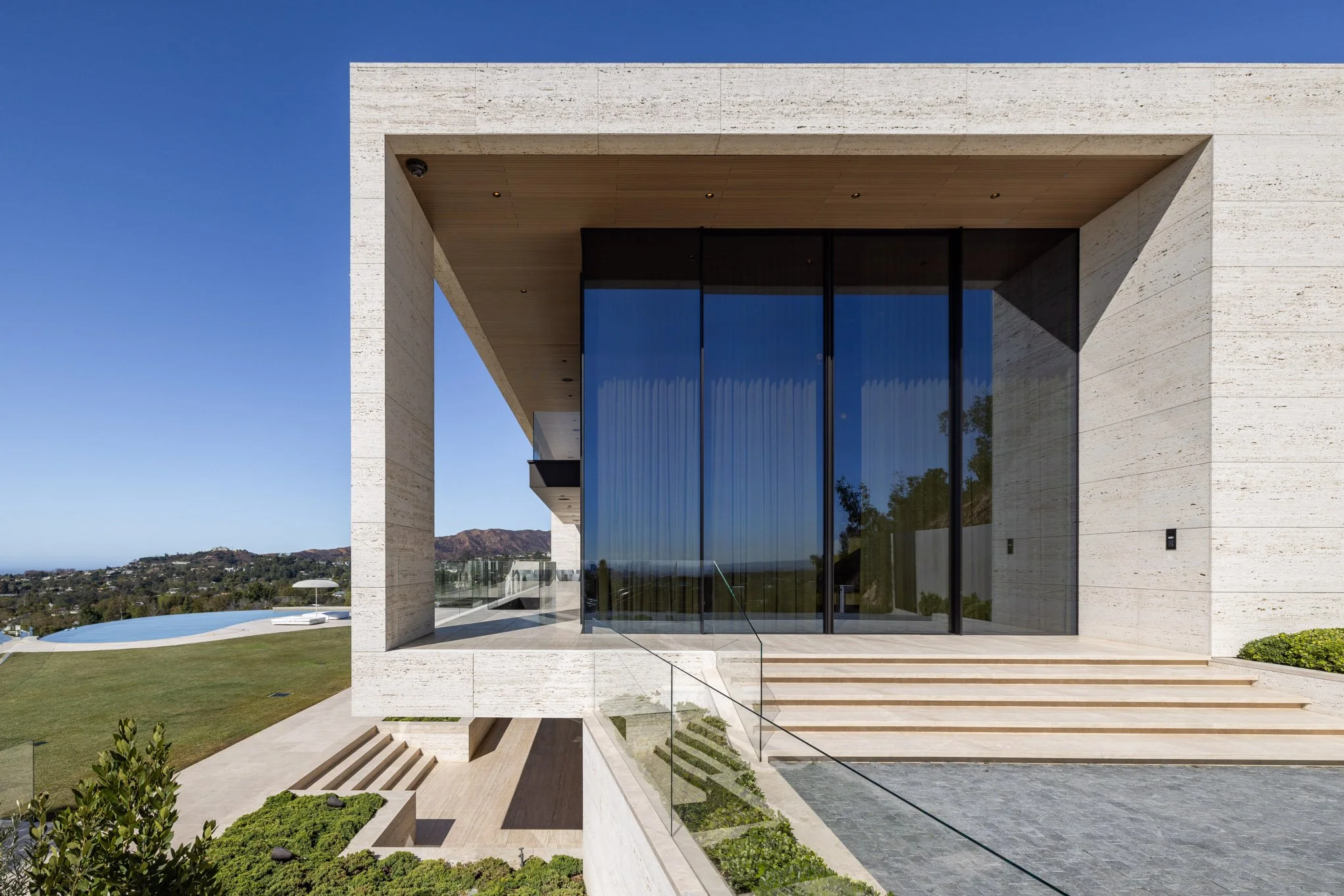 Modern house with large glass windows, concrete walls, and multiple steps leading to the entrance, overlooking a landscape with mountains and a pool in the distance.