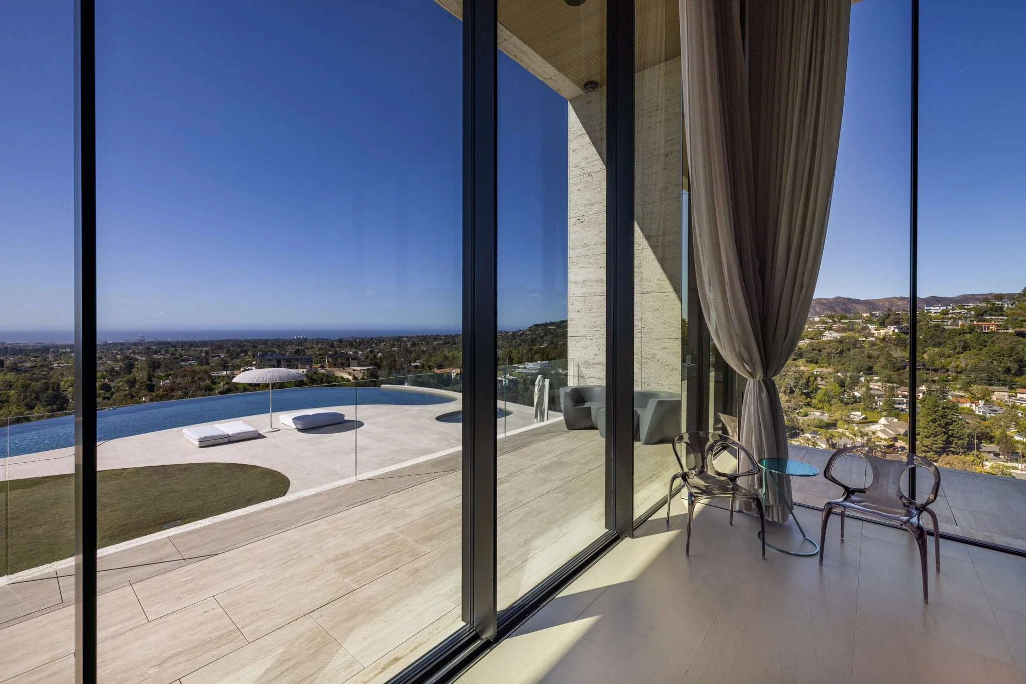 View of a modern balcony with glass doors, a swimming pool with lounge chairs and umbrella, overlooking a hilly cityscape and ocean in the distance under a clear blue sky.