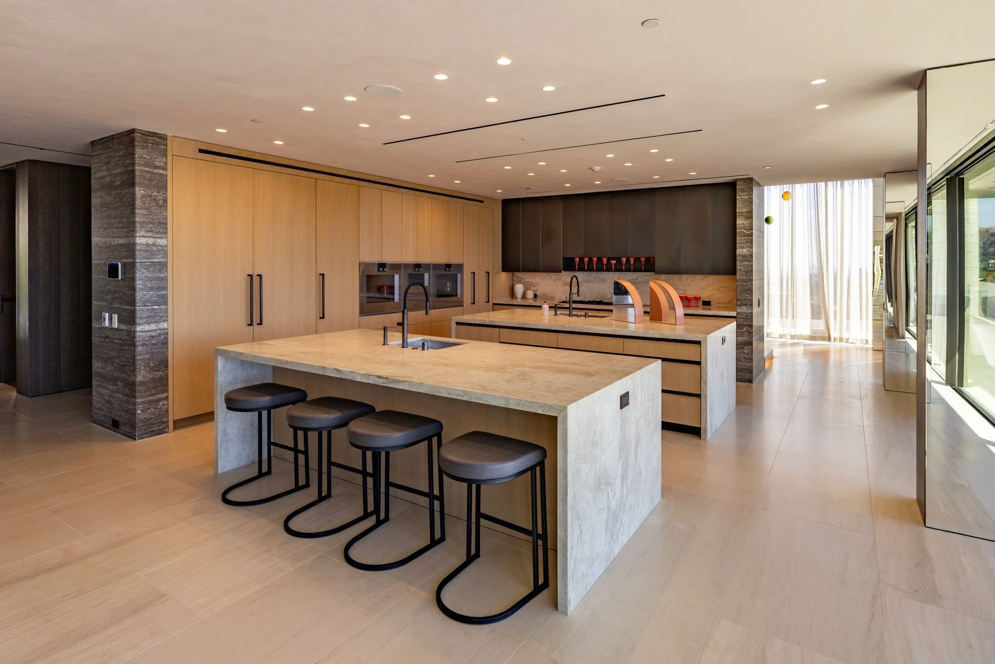 Modern kitchen with large marble island, light wood cabinetry, black appliances, and bar stools, with large windows and a sliding glass door leading outside.