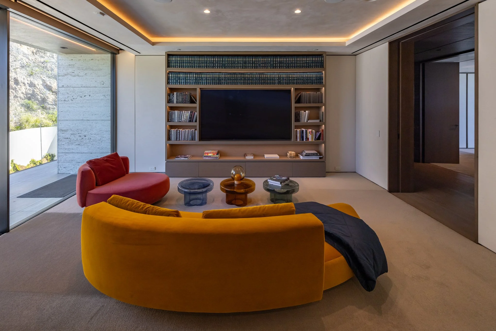 Modern living room with two colorful velvet sofas in yellow and red, glass tables, a large built-in bookshelf with books, a flat-screen TV, and a sliding glass door leading outside.