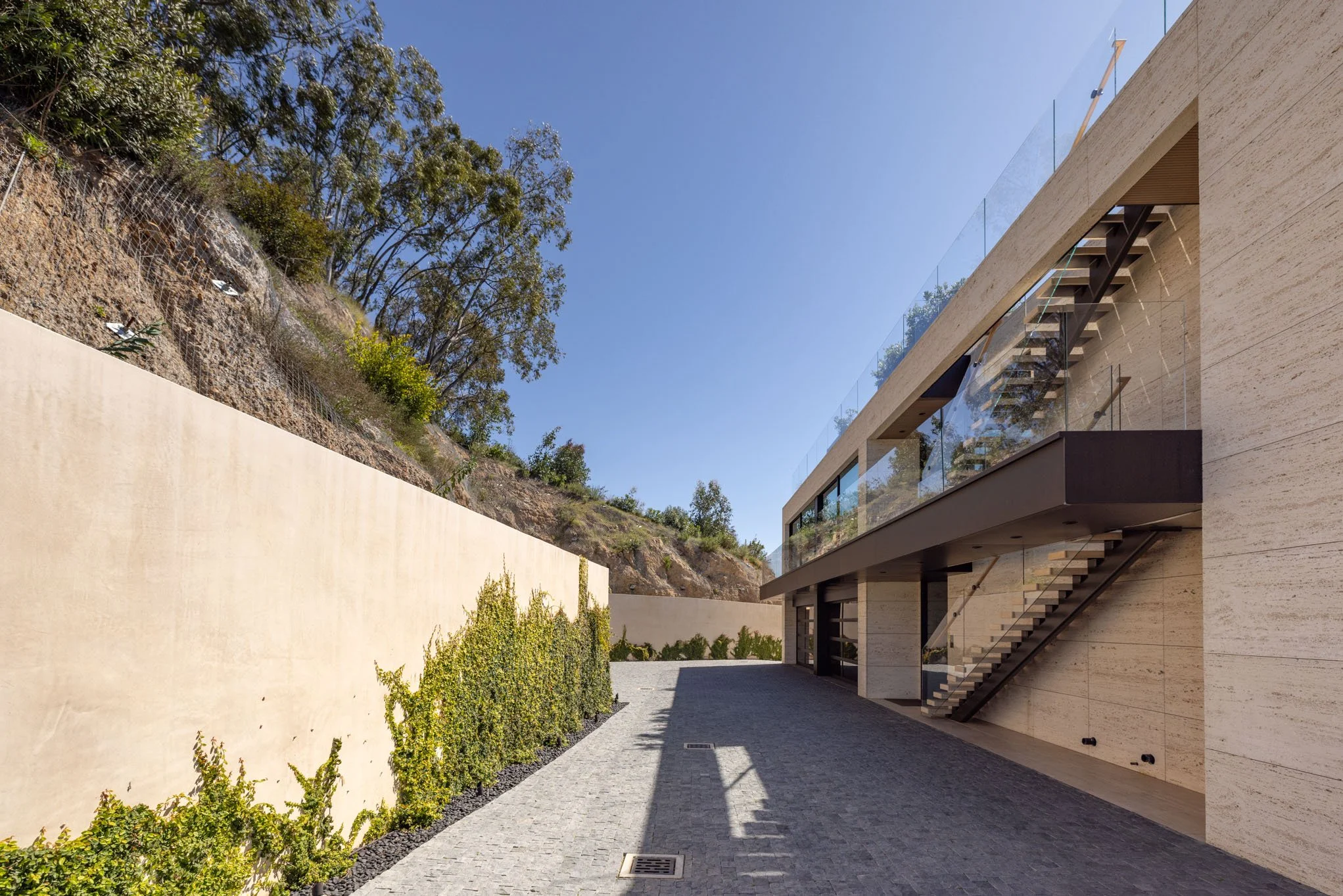 Modern building with glass balcony, outdoor staircase, paved driveway, and a landscape with trees and a hillside against a clear blue sky.