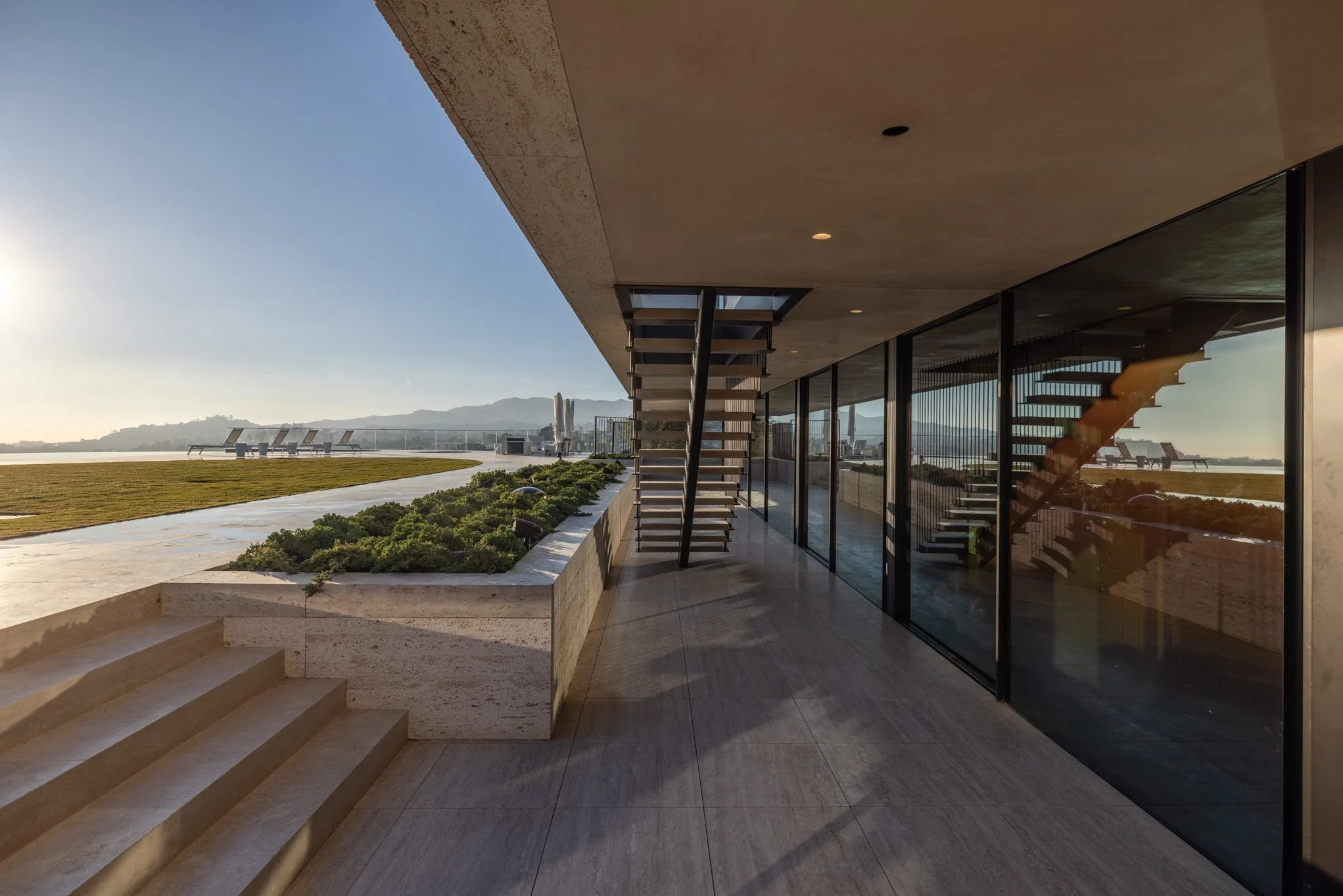 Modern building terrace with glass walls, outdoor stairs, landscaped planter, and a view of distant mountains and benches on the lawn connecting to the water.
