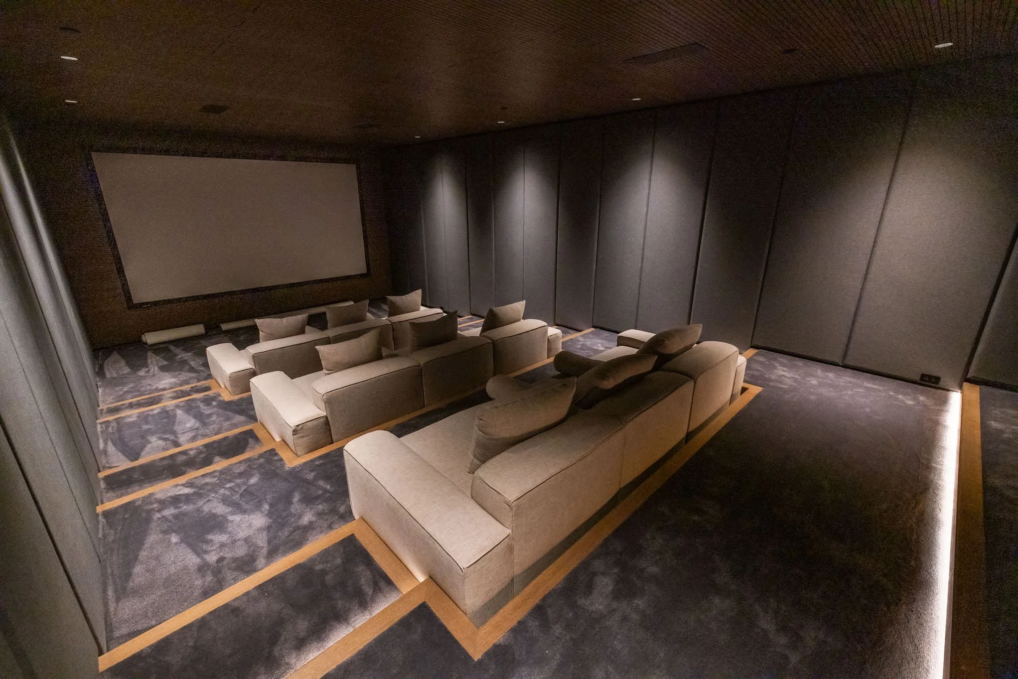Empty private movie theater with a large screen, beige sectional sofas, and dark walls.