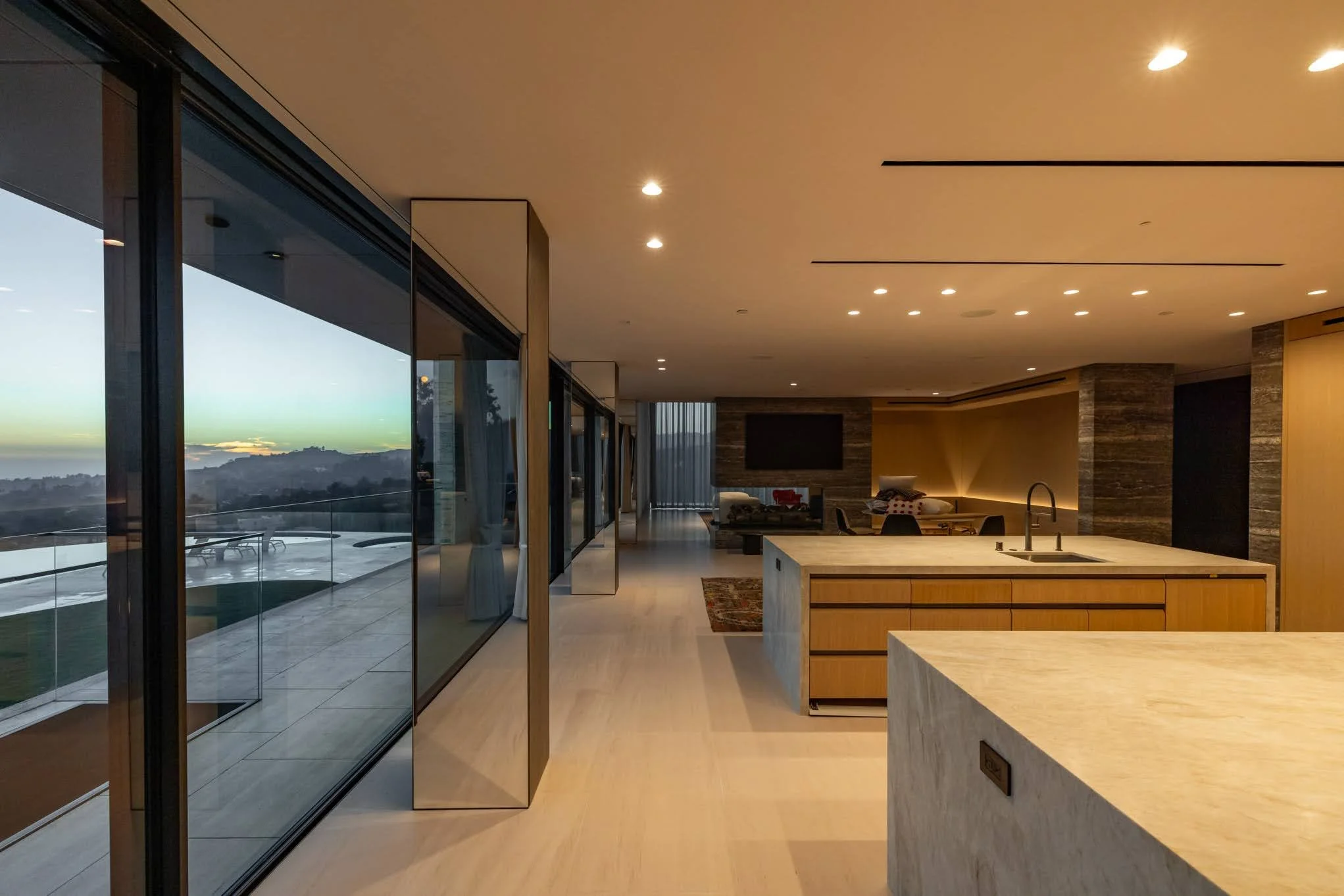 Modern open-concept living space with large glass sliding doors overlooking a sunset view, kitchen island with sink, and living area with television and seating.