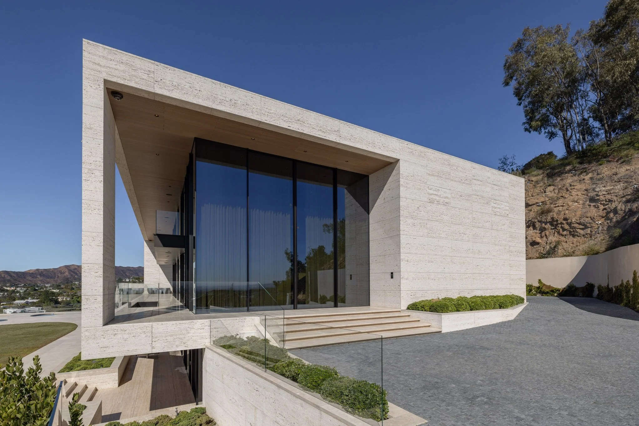 Modern building with large glass windows, concrete exterior, spiral staircase, and landscaped surroundings under a clear blue sky.