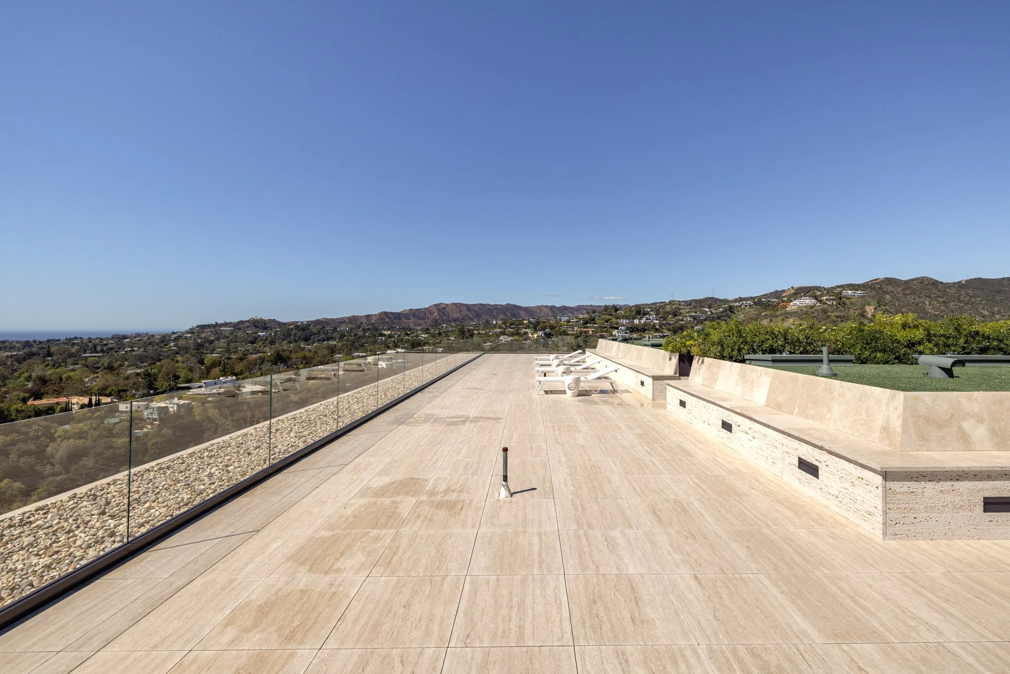 Rooftop terrace with beige tiles, glass railing, view of hilly landscape and blue sky in the background.