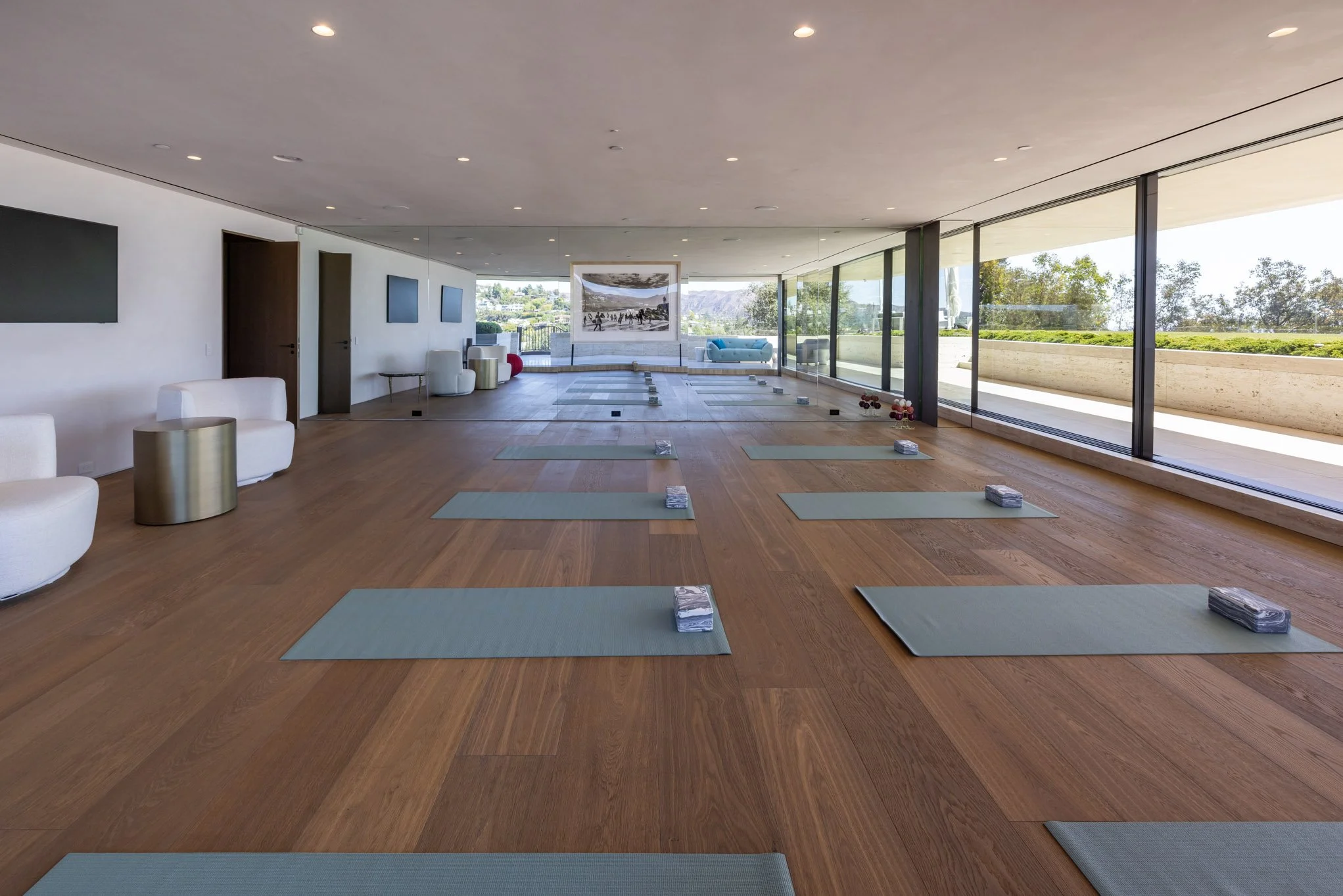 A spacious indoor yoga studio with wooden floors, large windows overlooking a garden, yoga mats and blocks arranged for a class, and modern white seating and wall-mounted TVs.
