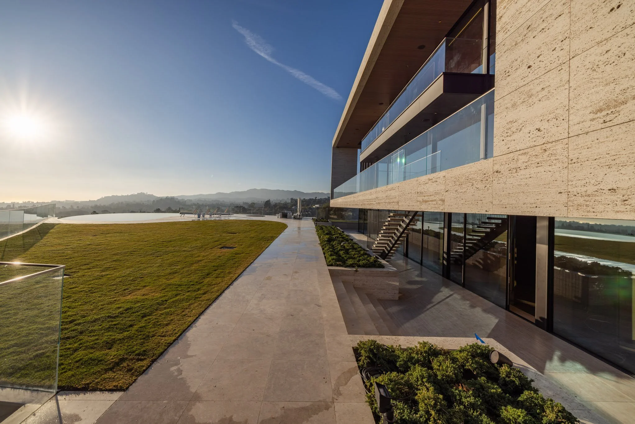 Modern building with glass balconies and stairs, outdoor patio with grassy lawn and mountain view in the distance, under clear sunny sky.