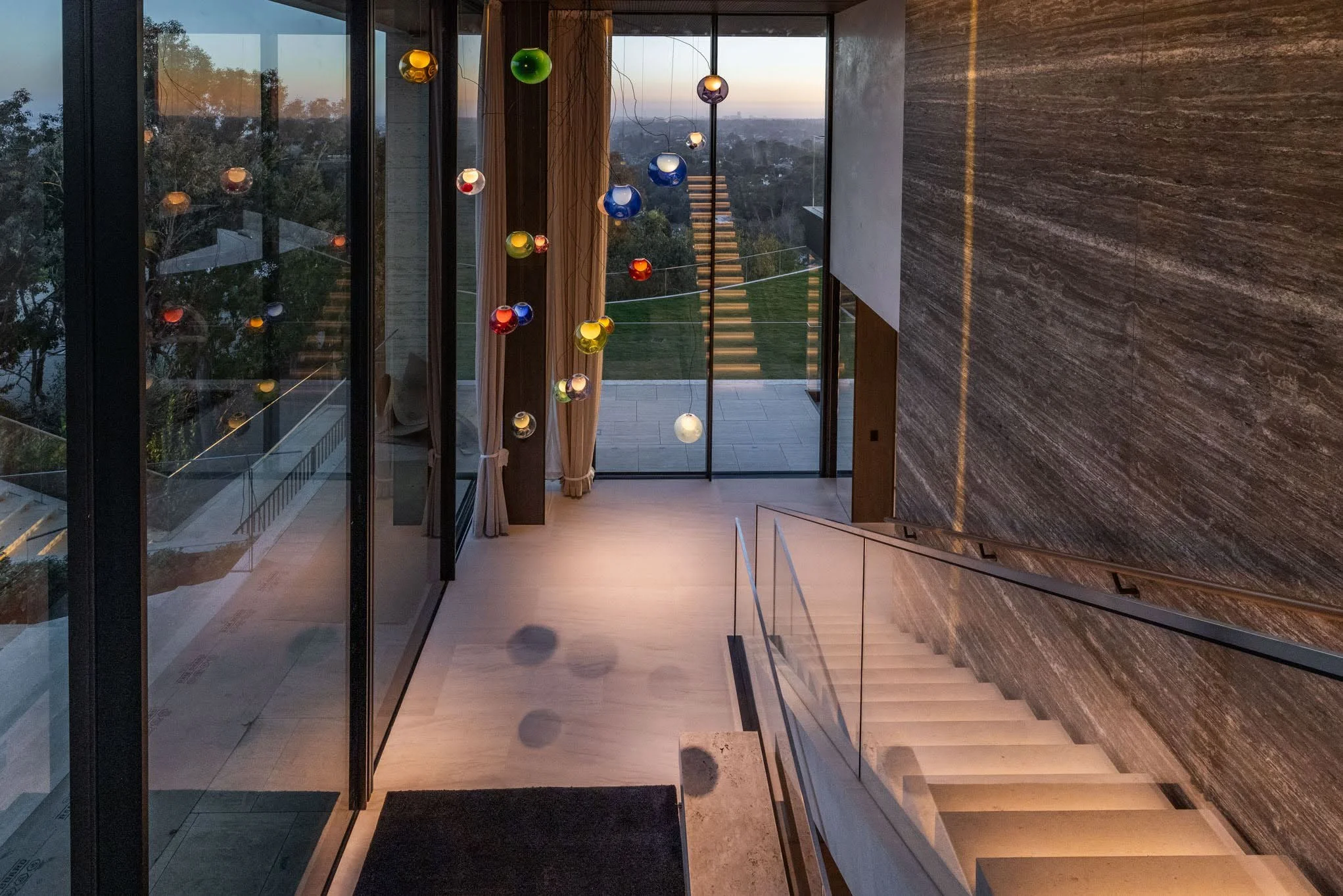 Interior view of a modern home with a staircase, large glass sliding doors leading to an outdoor patio, colorful pendant lights hanging from the ceiling, and a scenic view outside at sunset.
